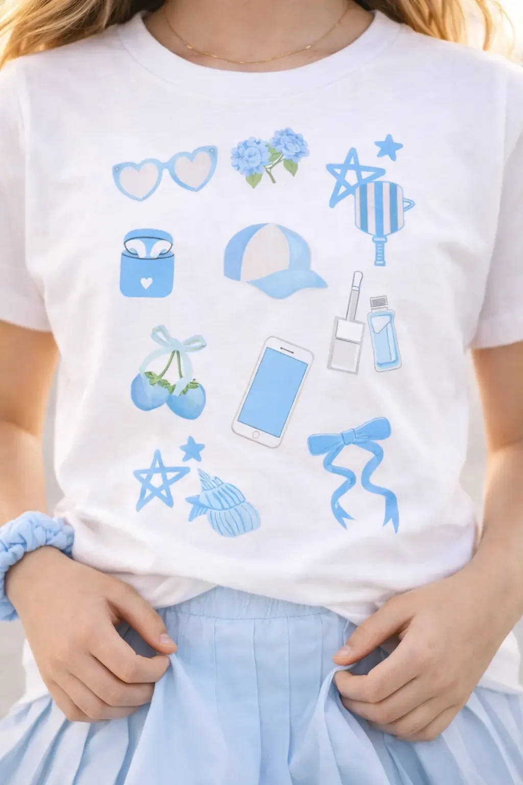 Close-up of a white graphic tee showing a collection of light blue preppy doodle icons including bows, a cap, and a phone.