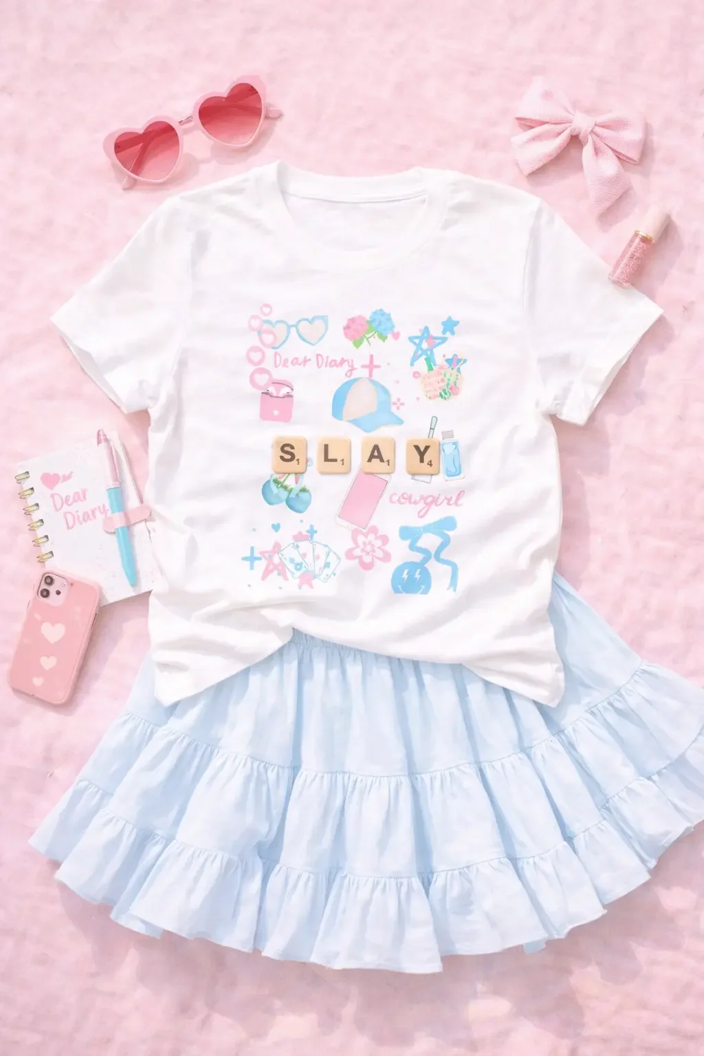 Flat lay of a white "SLAY" doodle icon graphic tee and a light blue ruffled tiered skort styled with heart sunglasses, a matching notebook, phone case, and hair bow.