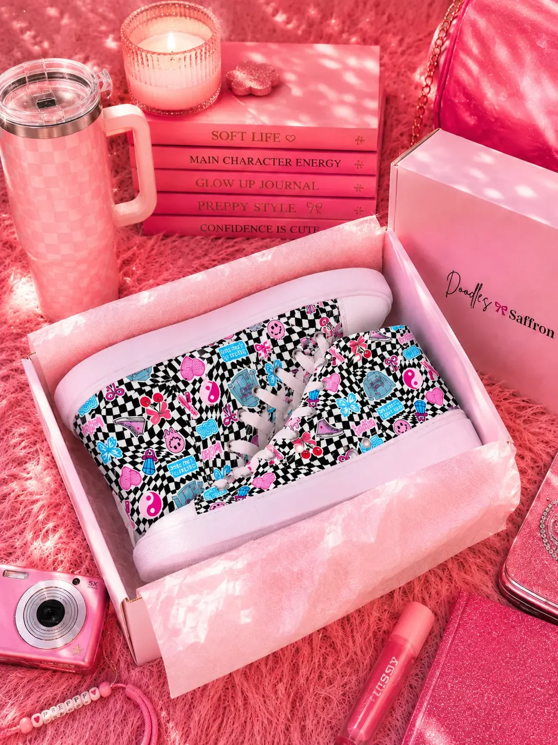 Black and white checkered high-top canvas sneakers inside a retail shipping box surrounded by pink preppy aesthetic accessories.