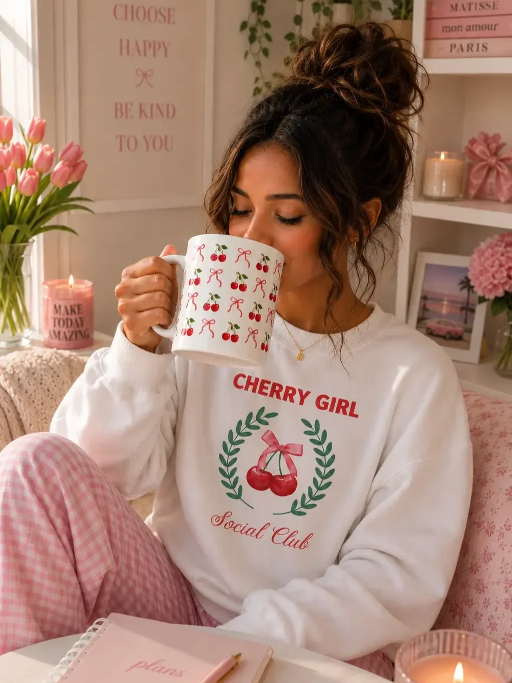 Woman in a white cherry girl sweatshirt drinking from a bow and cherry pattern mug while sitting at her desk.