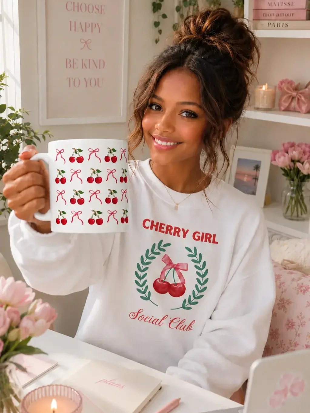 Smiling woman wearing a Cherry Girl Social Club sweatshirt holds up a white mug patterned with bows and cherries.