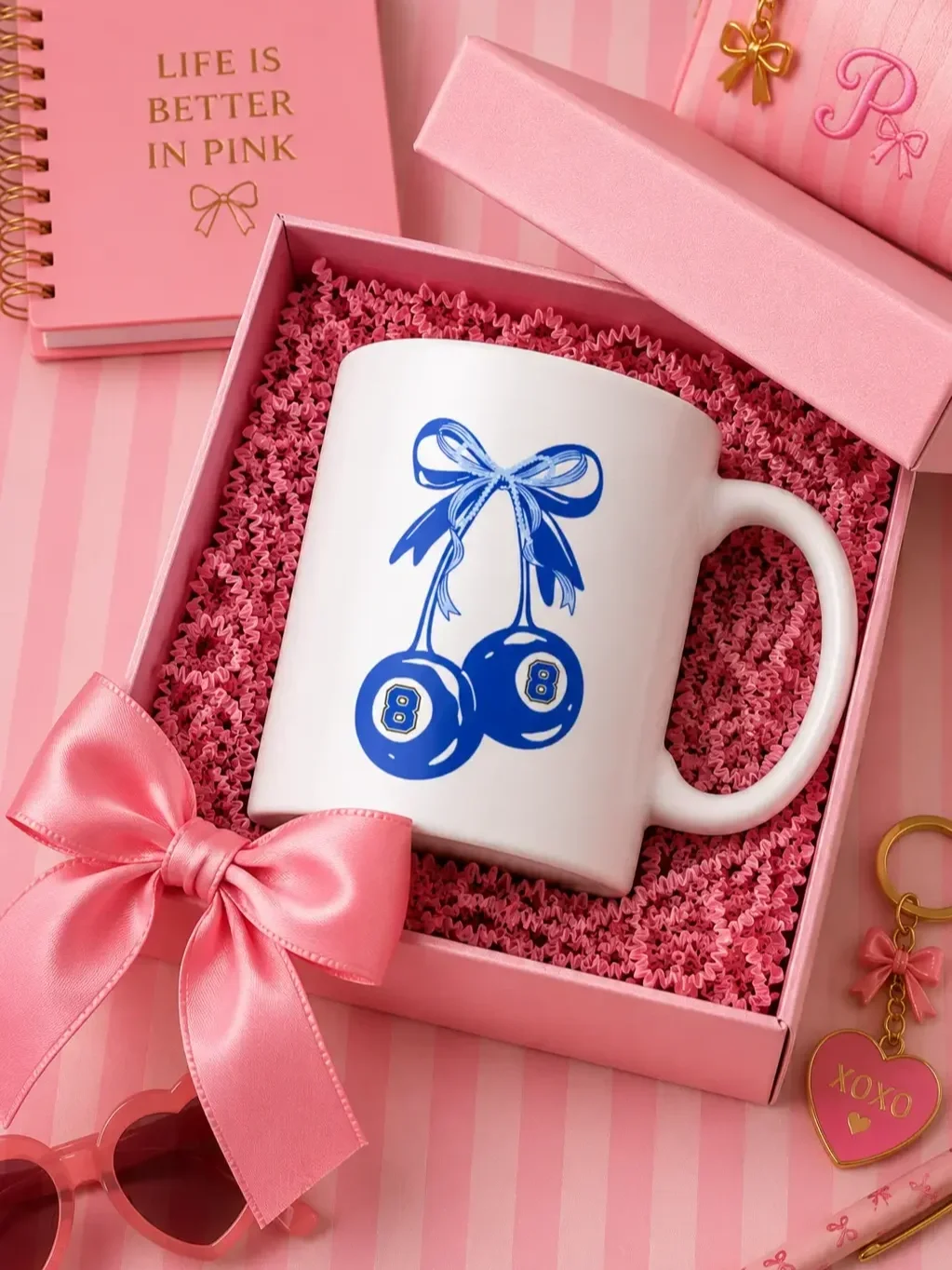 White mug featuring a blue eight-ball cherry design inside a pink gift box next to a pink notebook and stationary.