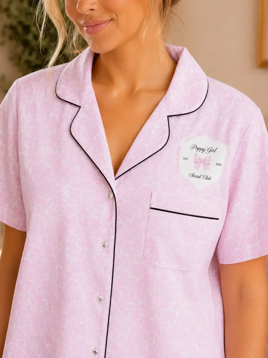 Close-up of a light pink patterned button-up pajama shirt featuring black piping details and a Preppy Girl Social Club crest design on the chest.
