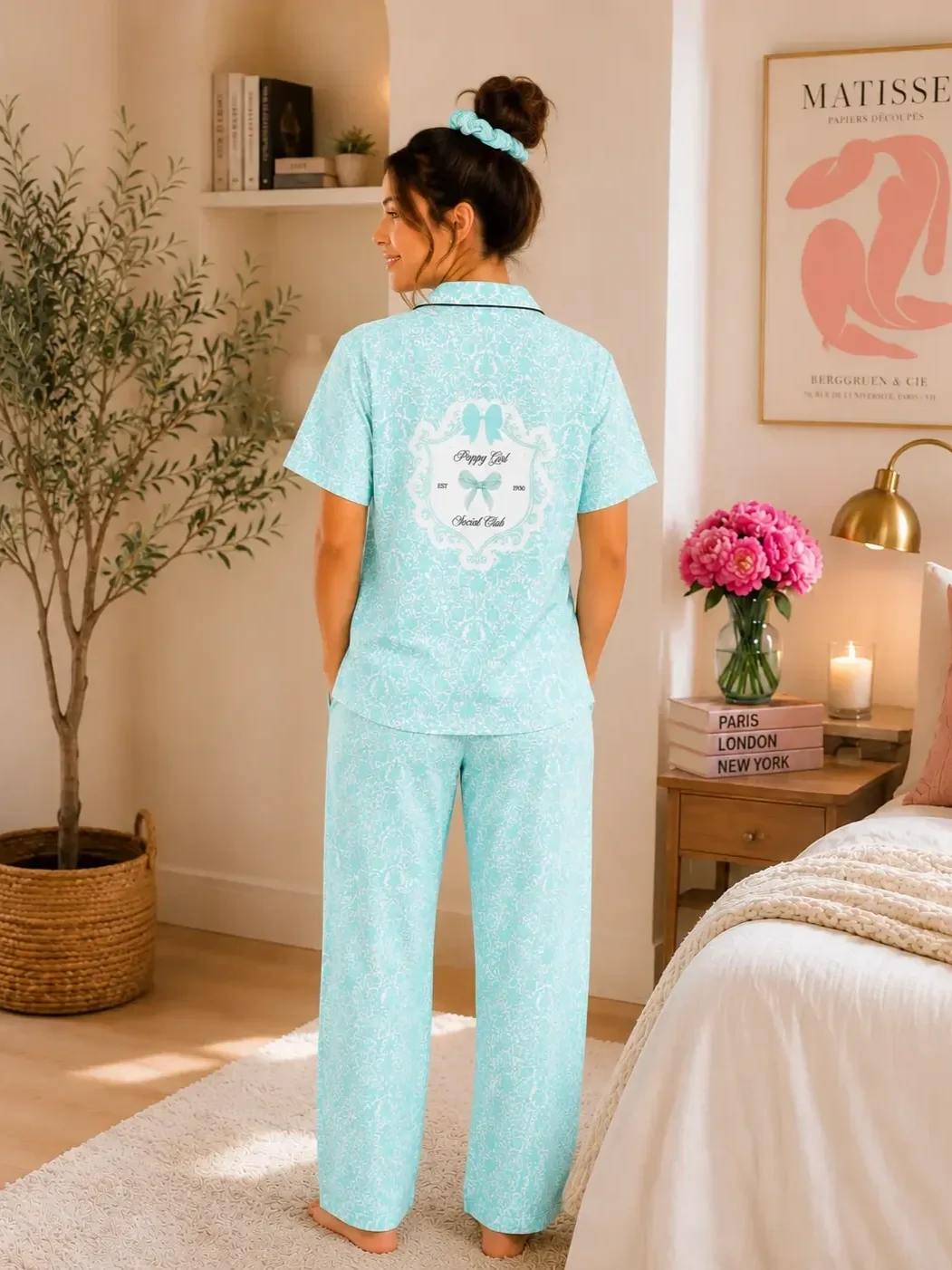Back view of a young woman standing in a bedroom wearing a matching mint green patterned pajama set with Preppy Girl Social Club graphic text on the shirt back.