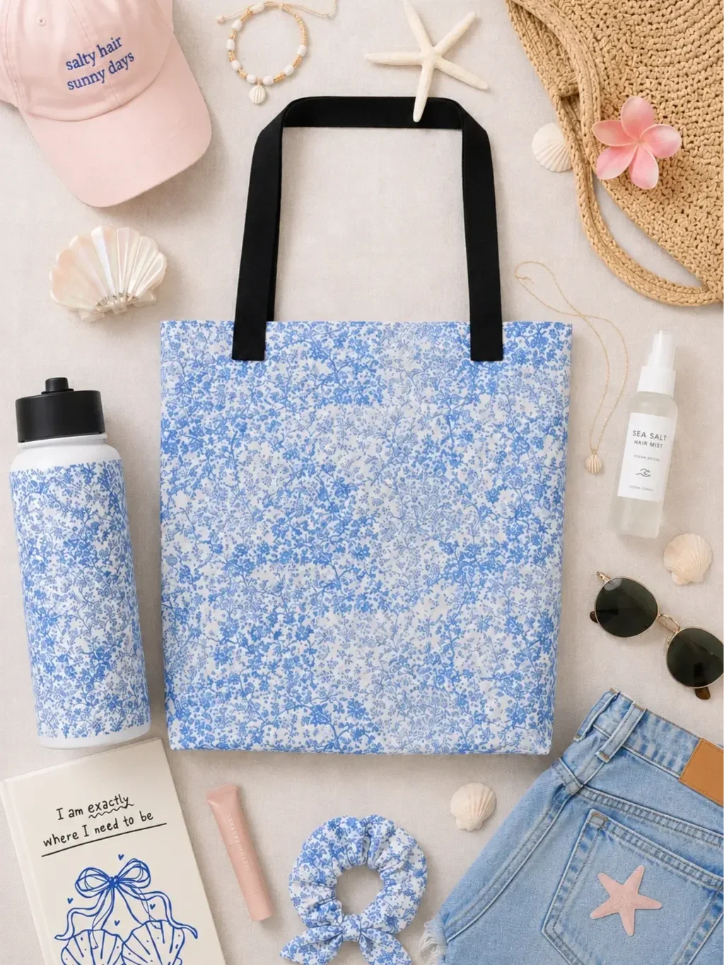 Blue floral tote bag flat lay with beach essentials like a pink cap, denim shorts, sunglasses, and a novel.
