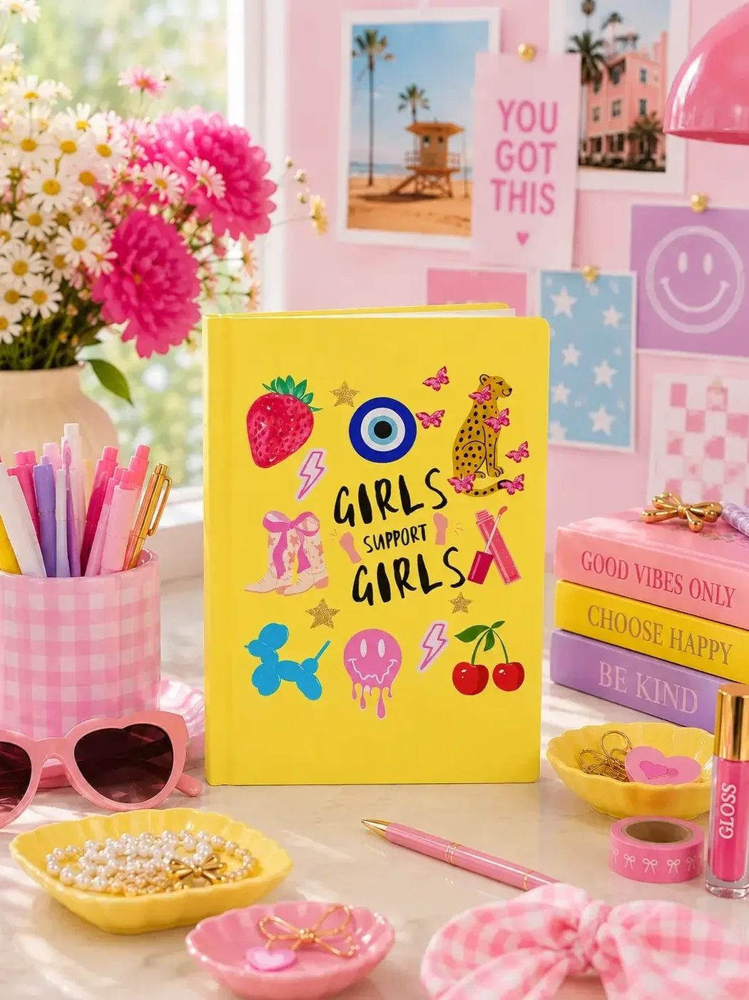 Bright yellow "Girls Support Girls" journal featuring coquette doodles, styled on an aesthetic pink desk setup with flowers.