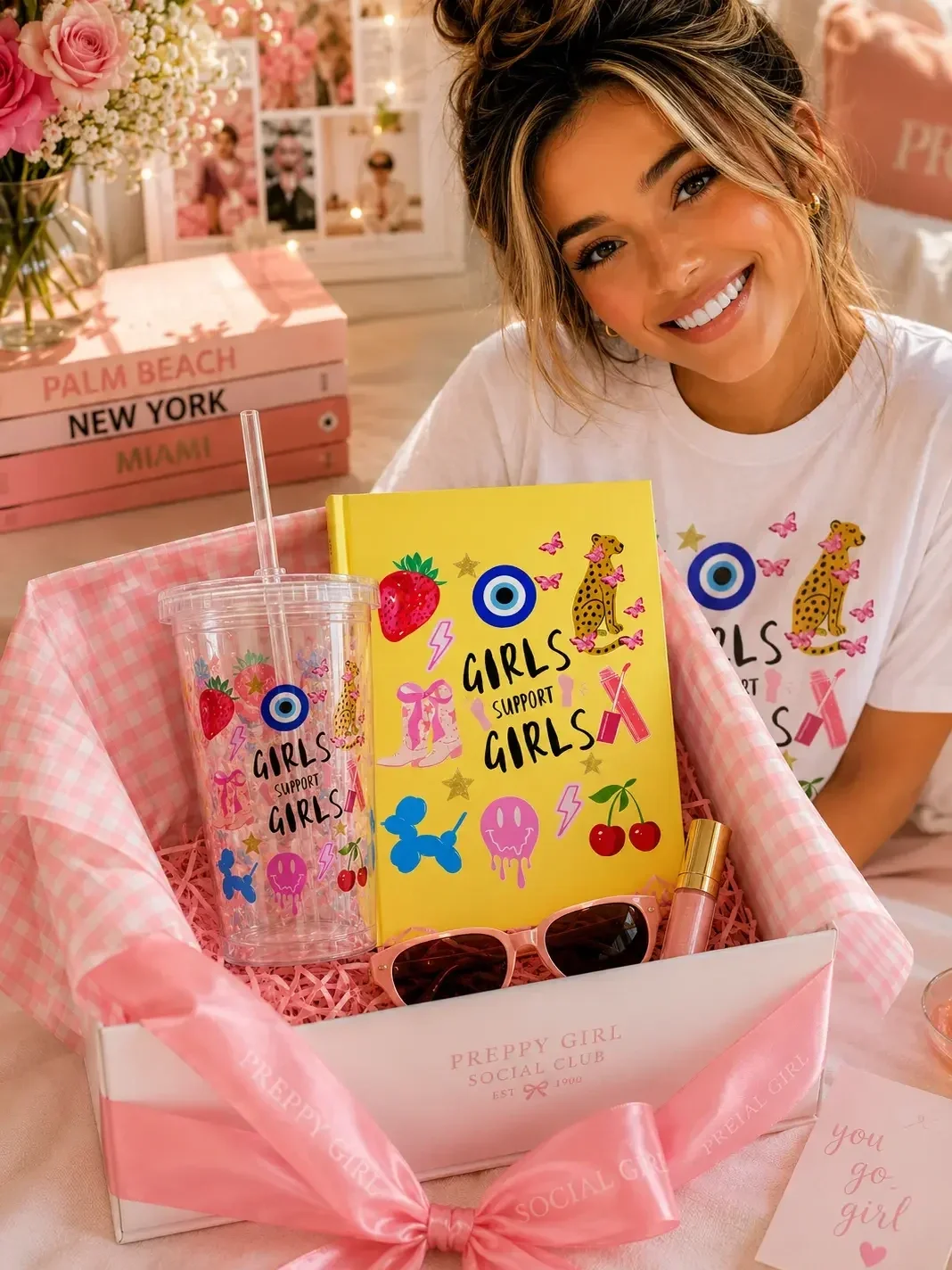 Smiling woman with a hair bun sitting behind a pink preppy gift box filled with a yellow Girls Support Girls journal and matching tumbler.