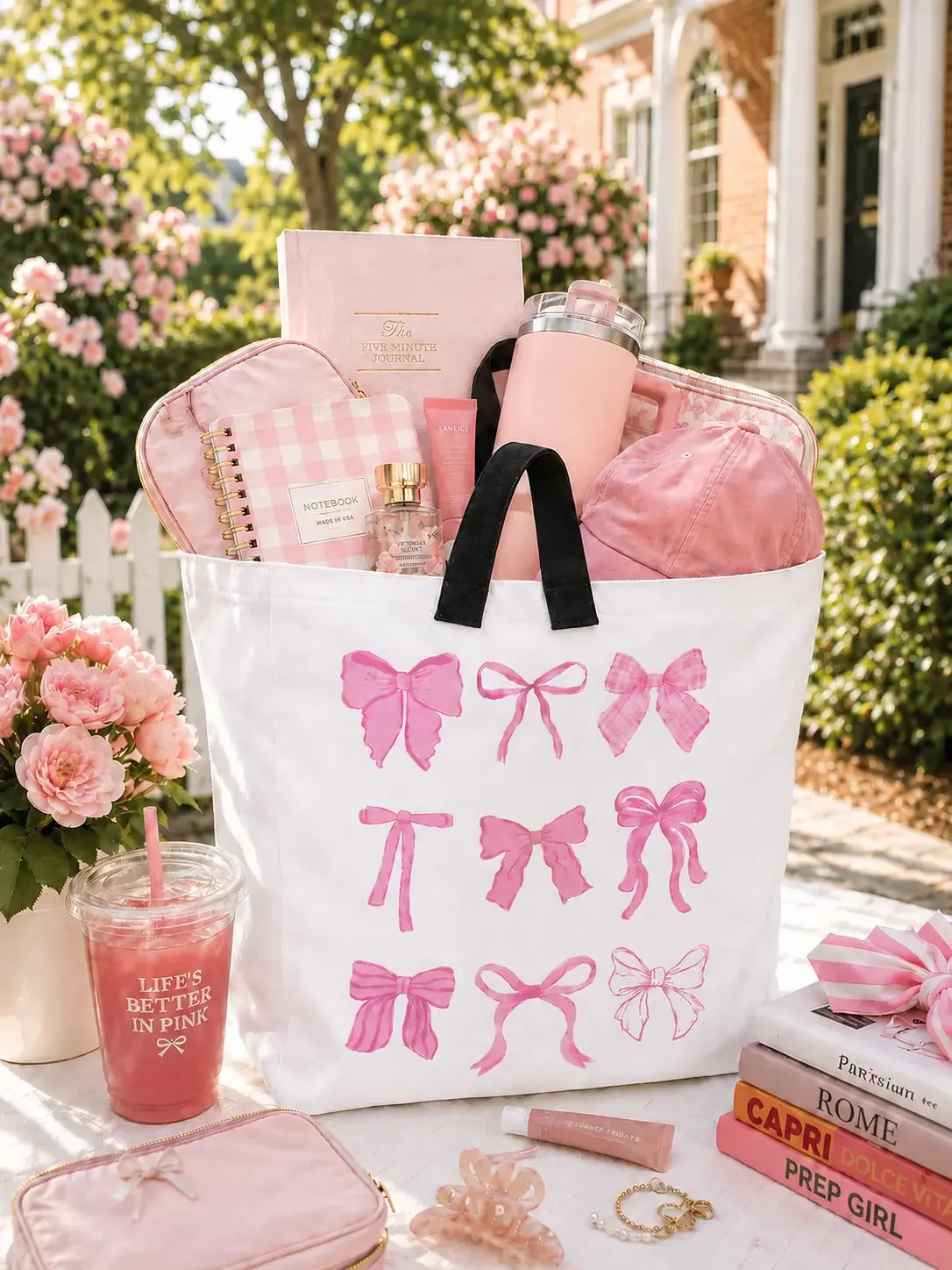 White ribbon bow tote bag filled with pink books, stationery, a pink Stanley tumbler, and a cap on a decorated desk setup.