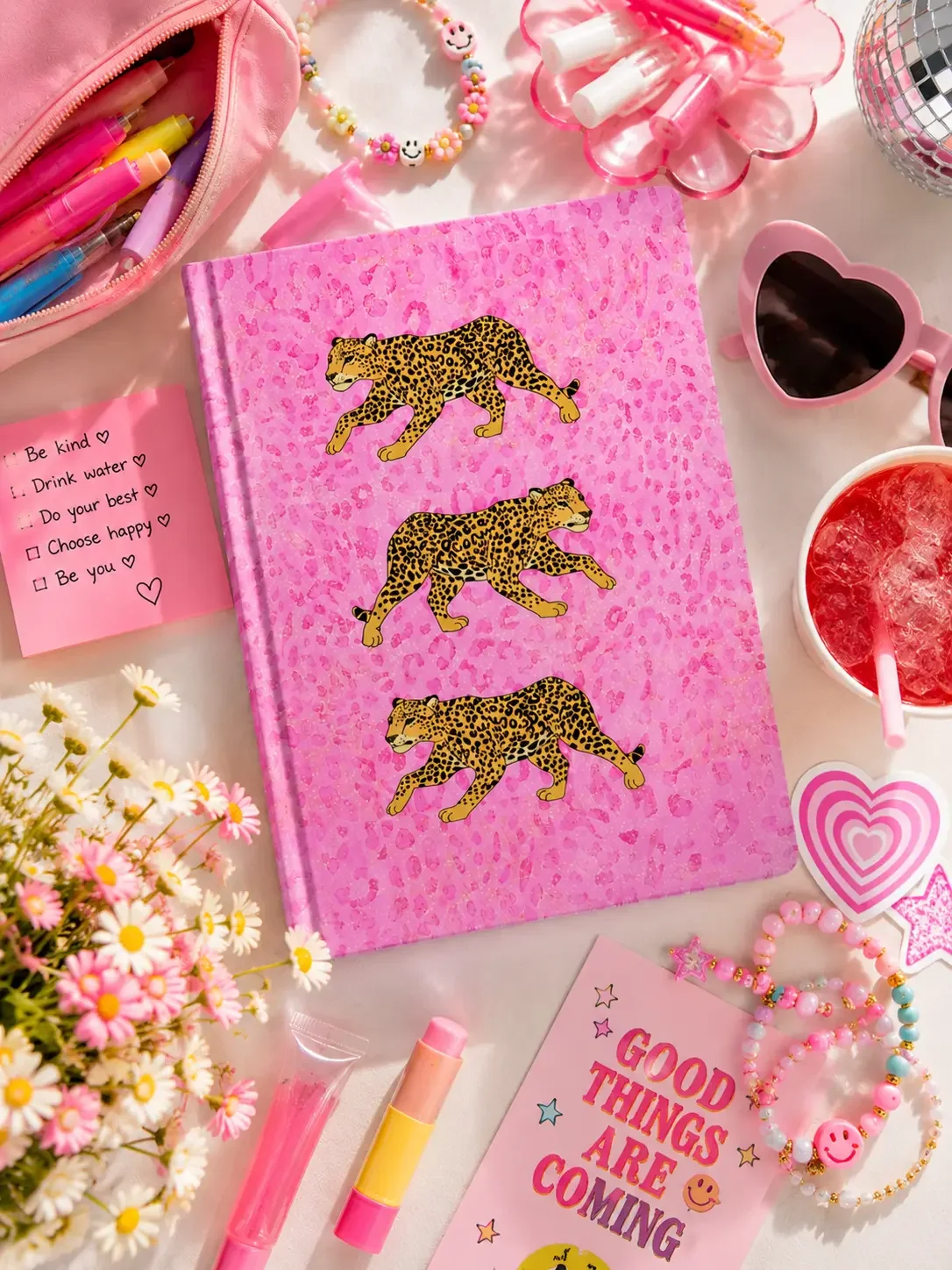 Flat lay of a pink leopard print journal with three cheetah illustrations, surrounded by pink heart sunglasses, flowers, and lip gloss.