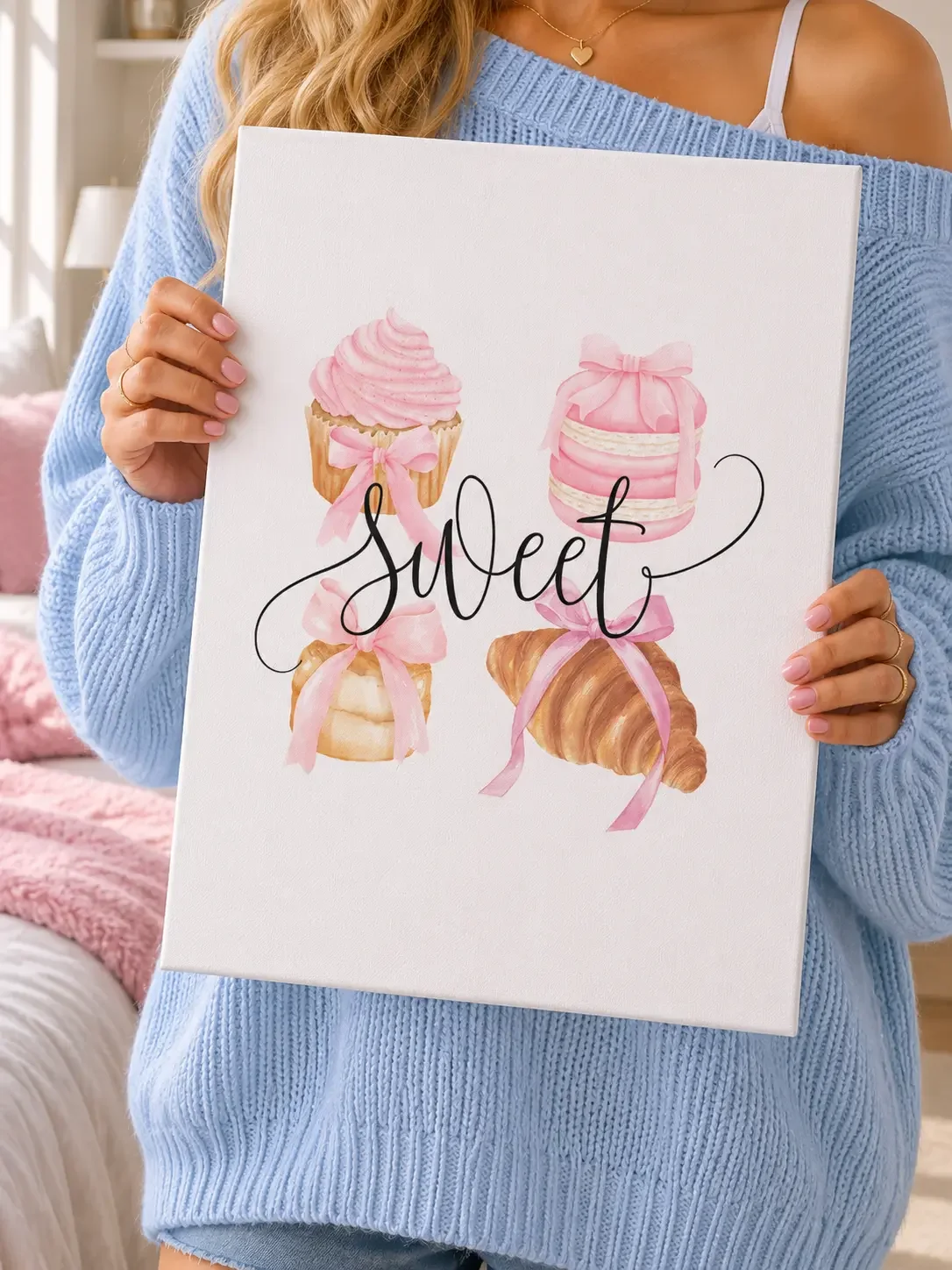 Girl in a light blue sweater holding a "Sweet" canvas art print featuring pink bow pastries and a croissant.
