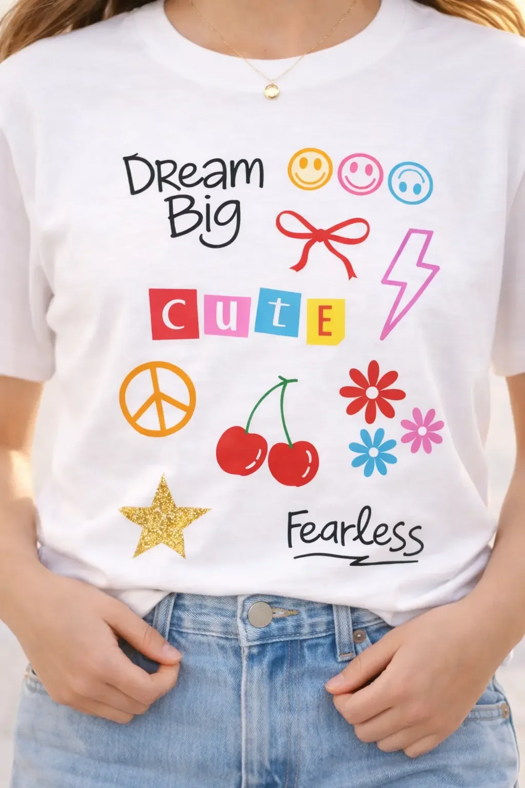Close-up of a white graphic tee showing preppy doodles like cherries, a coquette ribbon bow, smiley faces, and text reading "Dream Big" and "Fearless".