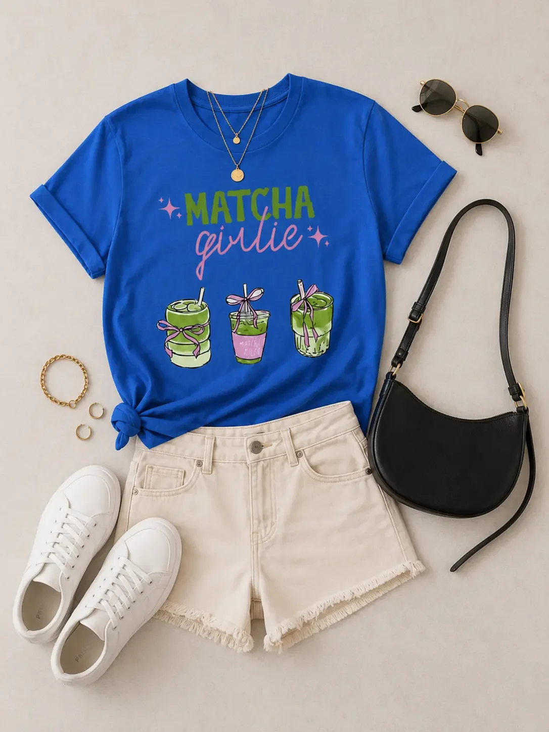 Flat lay display of a blue Matcha Girlie graphic t-shirt, cream denim shorts, white sneakers, a black shoulder bag, and sunglasses.