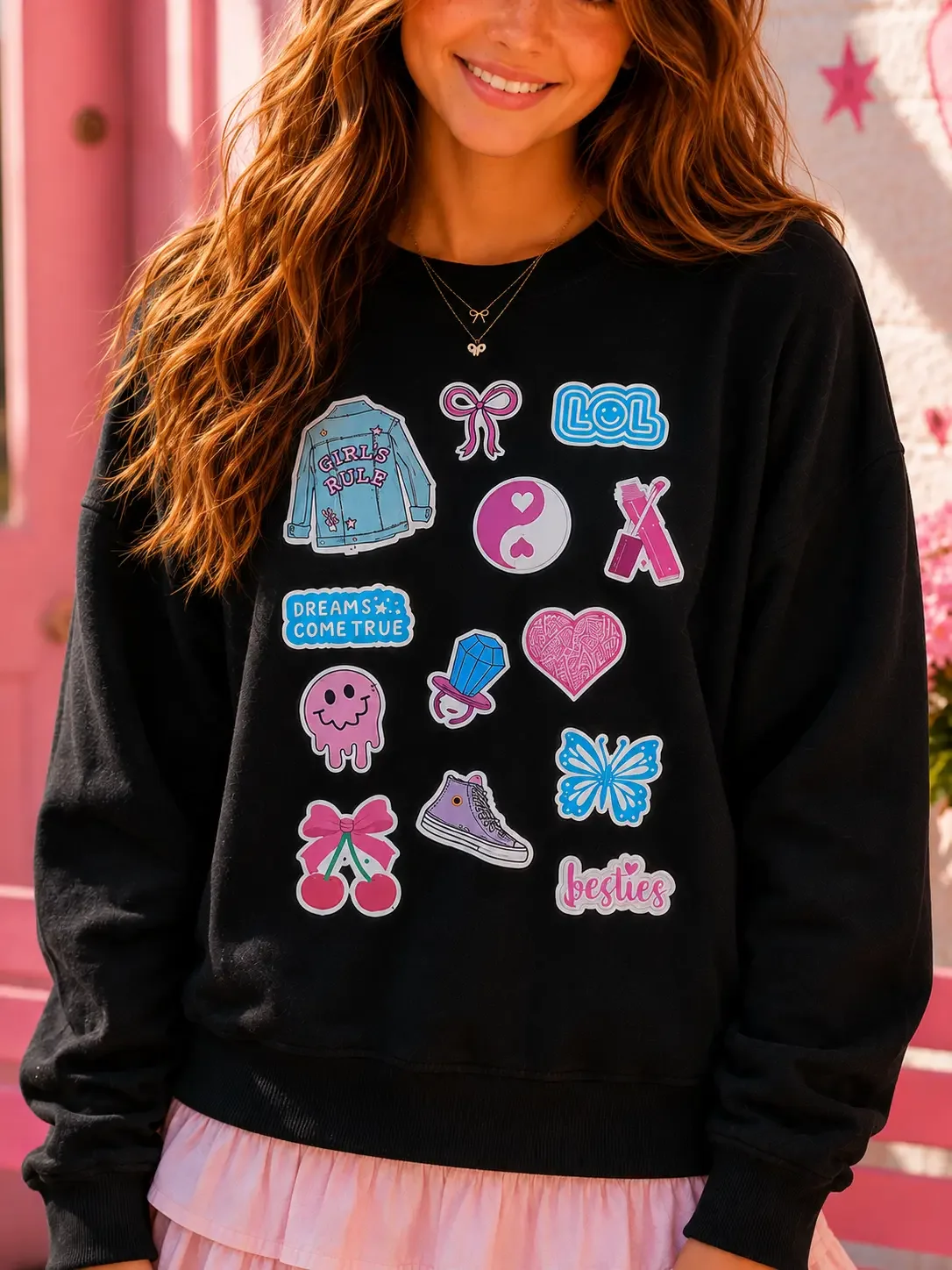 Close-up of a woman in a black crewneck sweatshirt covered in colorful retro sticker graphics, showing a peek of a pink ruffle skirt and layered necklaces.