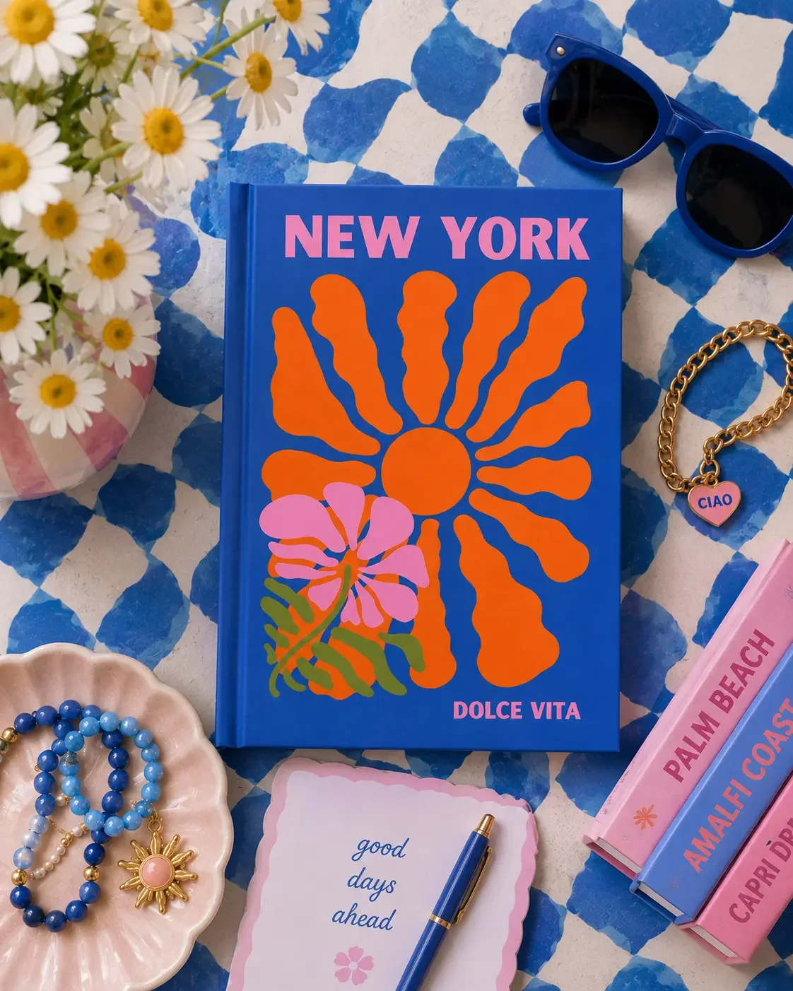 Bright blue "New York Dolce Vita" journal with orange flower graphic on a blue checkered background with stacked books and daisies.