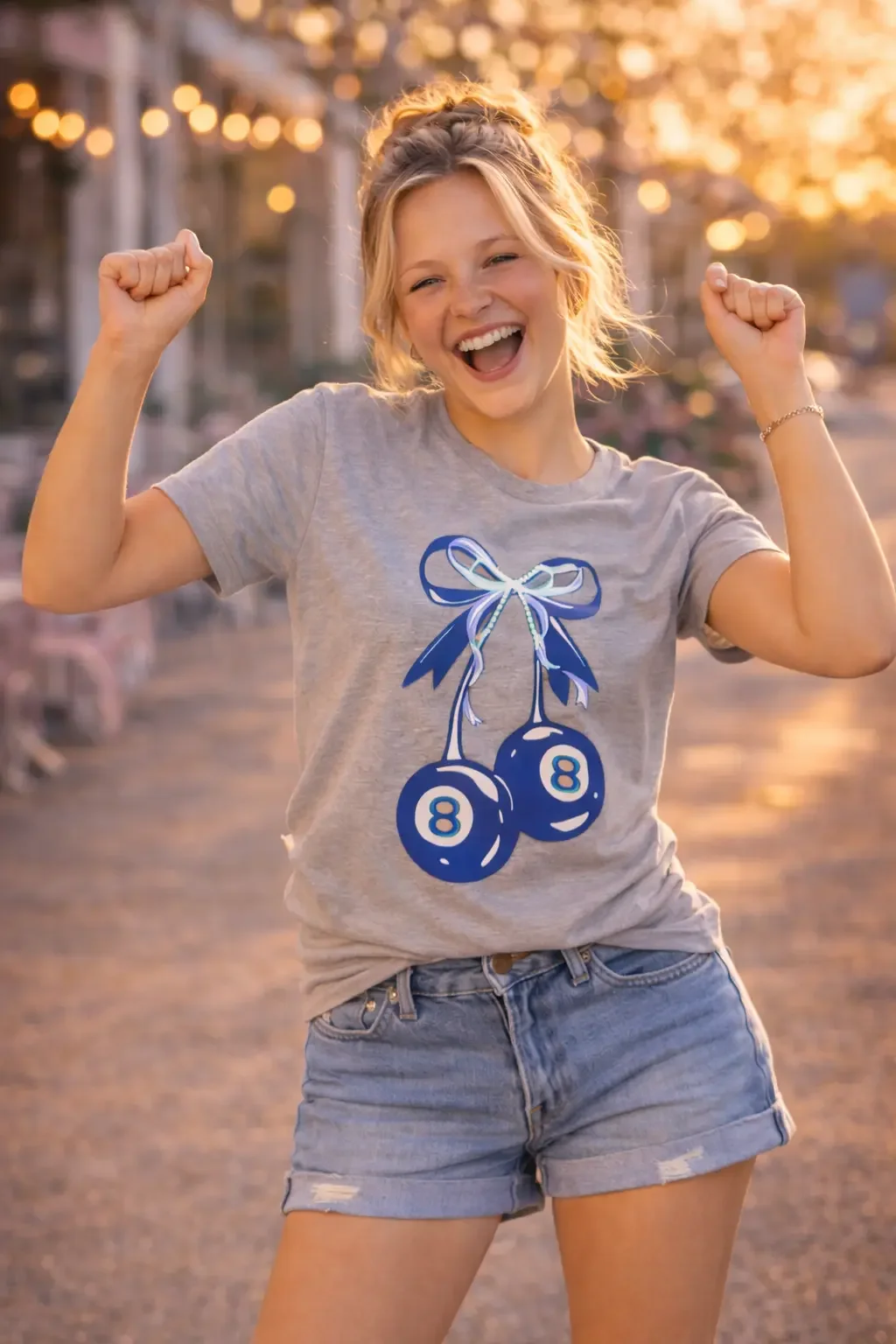 Cheerful girl posing excitedly with raised fists on a street wearing a grey blue-cherry graphic tee and denim shorts.