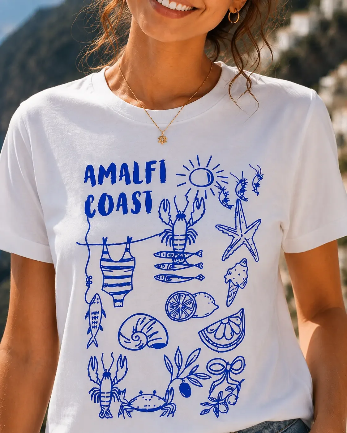 Close-up view of a white crew neck t-shirt featuring blue Amalfi Coast text above Mediterranean doodle graphic illustrations.