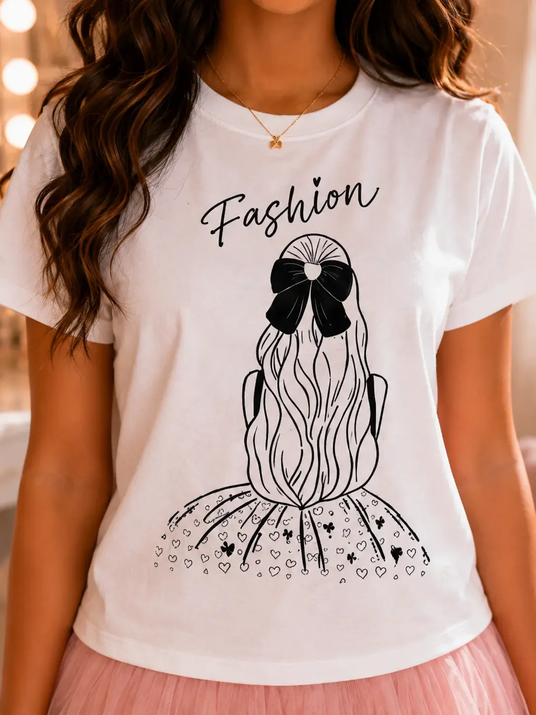 Close-up of a white graphic tee showing cursive "Fashion" text above a fine-line drawing of the back of a girl wearing a large black hair bow and a skirt filled with heart patterns.