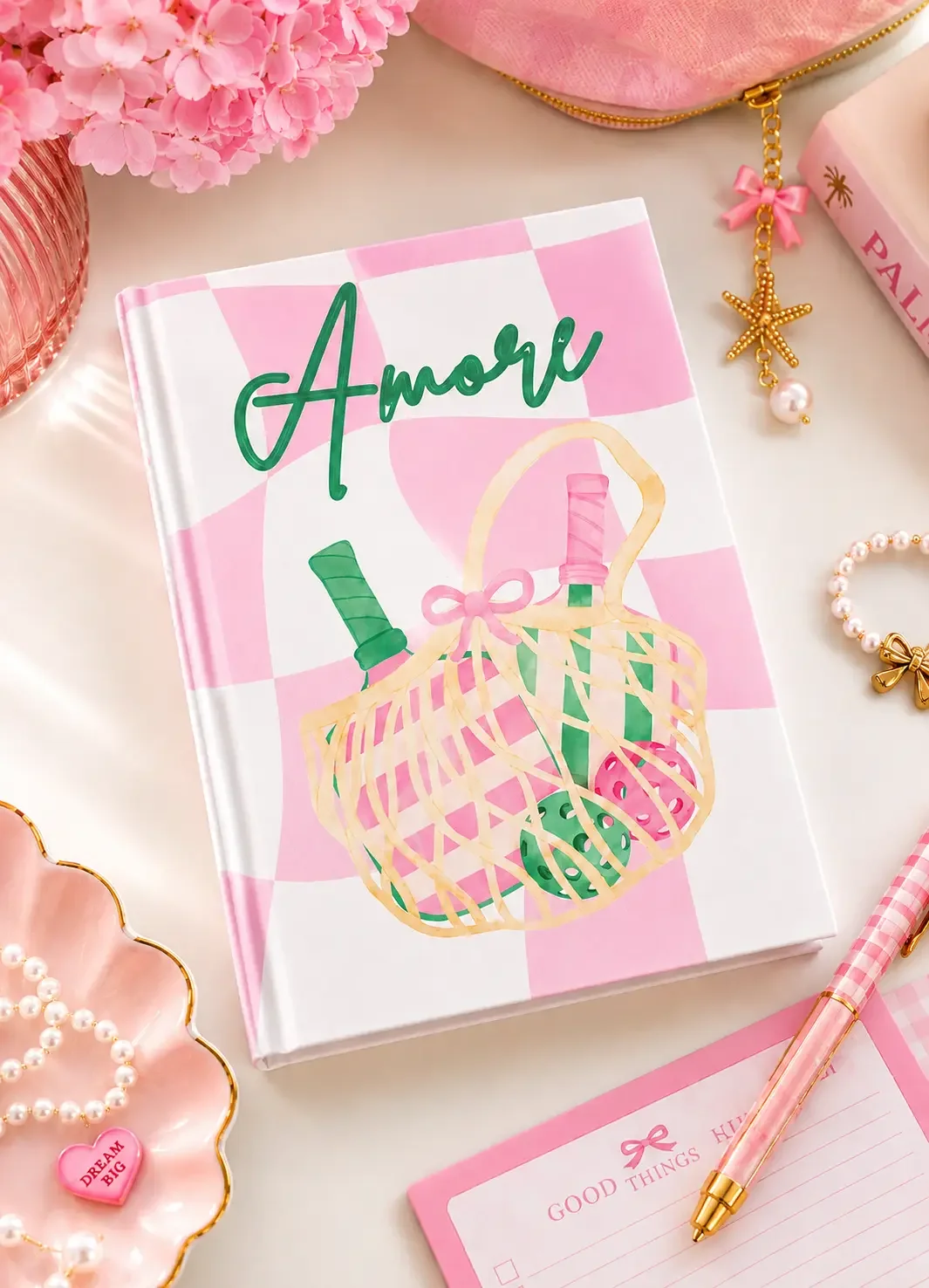 Flat lay of a pink checkered "Amore" pickleball notebook on a pink desk with a matching pink pen, pearls, and hydrangeas.