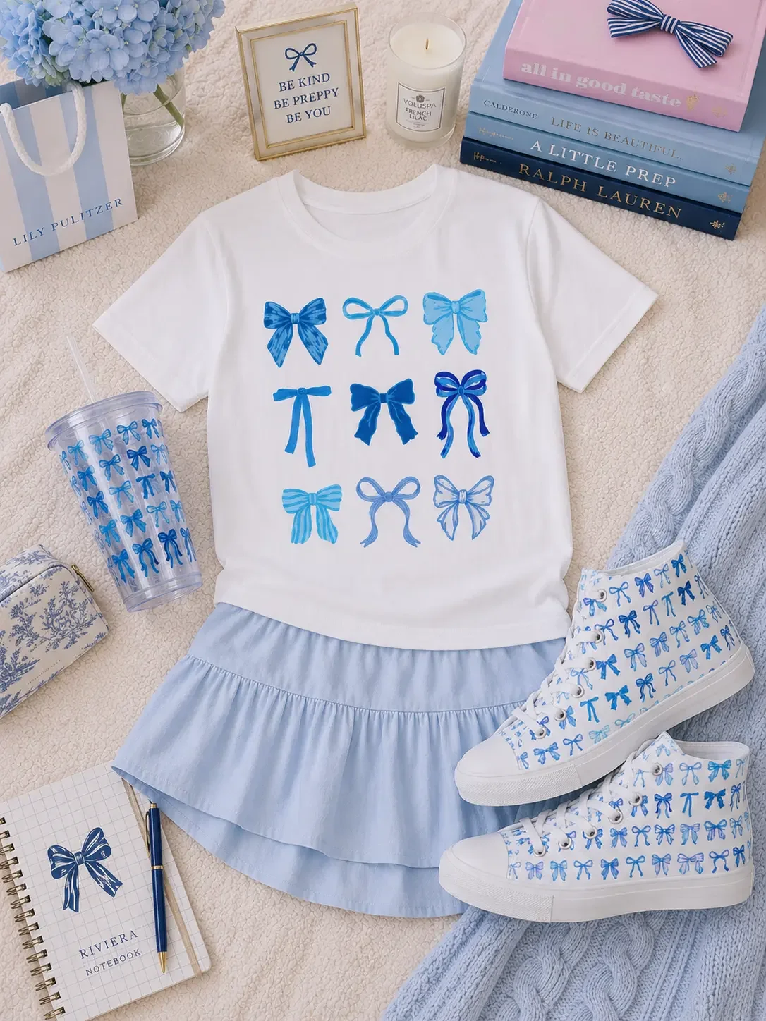 Flat lay of a white blue-bow graphic tee, light blue skirt, matching high-top sneakers, a tumbler, and notebooks.
