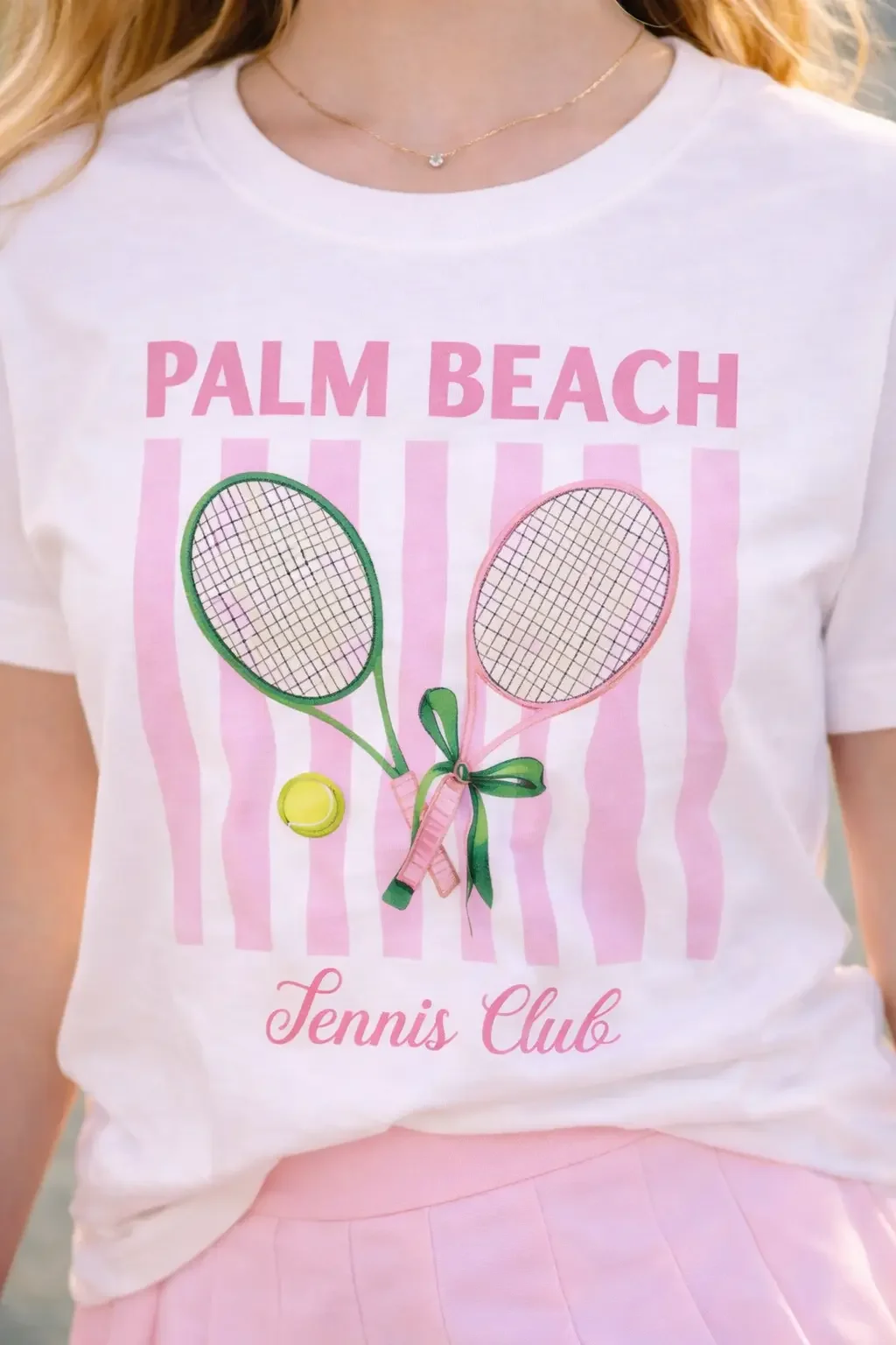 Close up of a white graphic t-shirt featuring pink stripes with vintage tennis rackets and text tucked into a pink pleated skirt.