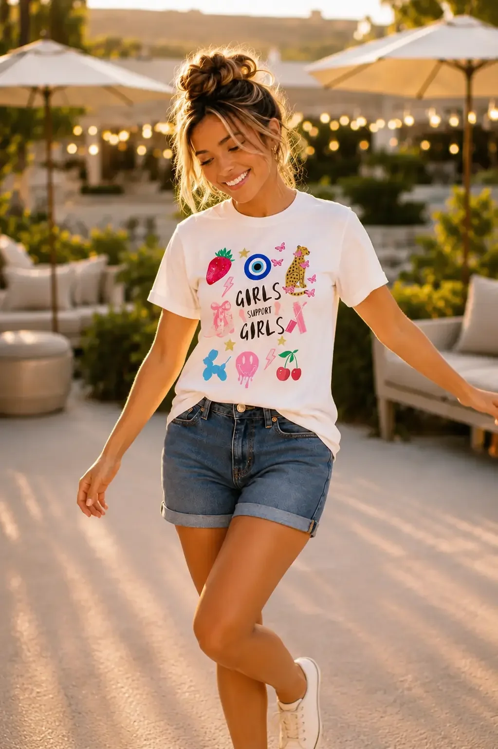 Smiling woman walking in a white Girls Support Girls graphic t-shirt, cuffed denim shorts, and white sneakers.
