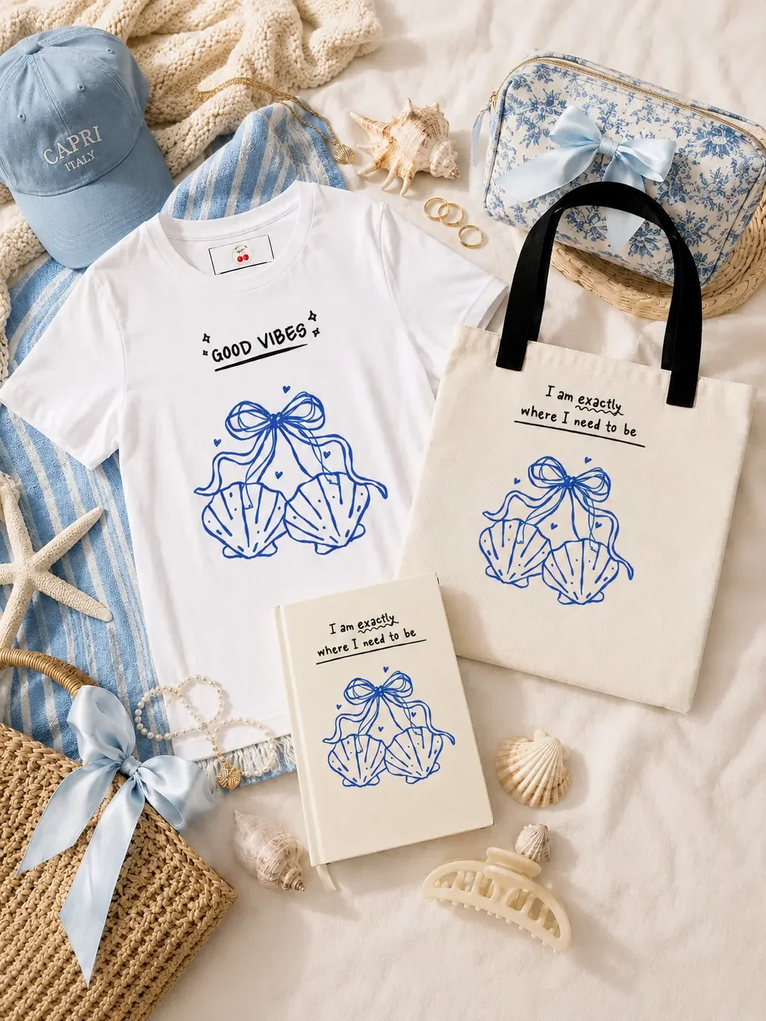 Flat lay display of a white graphic t-shirt, matching canvas tote bag, and a hardcover journal with blue seashell bow graphics, styled with beach accessories. (Copy)