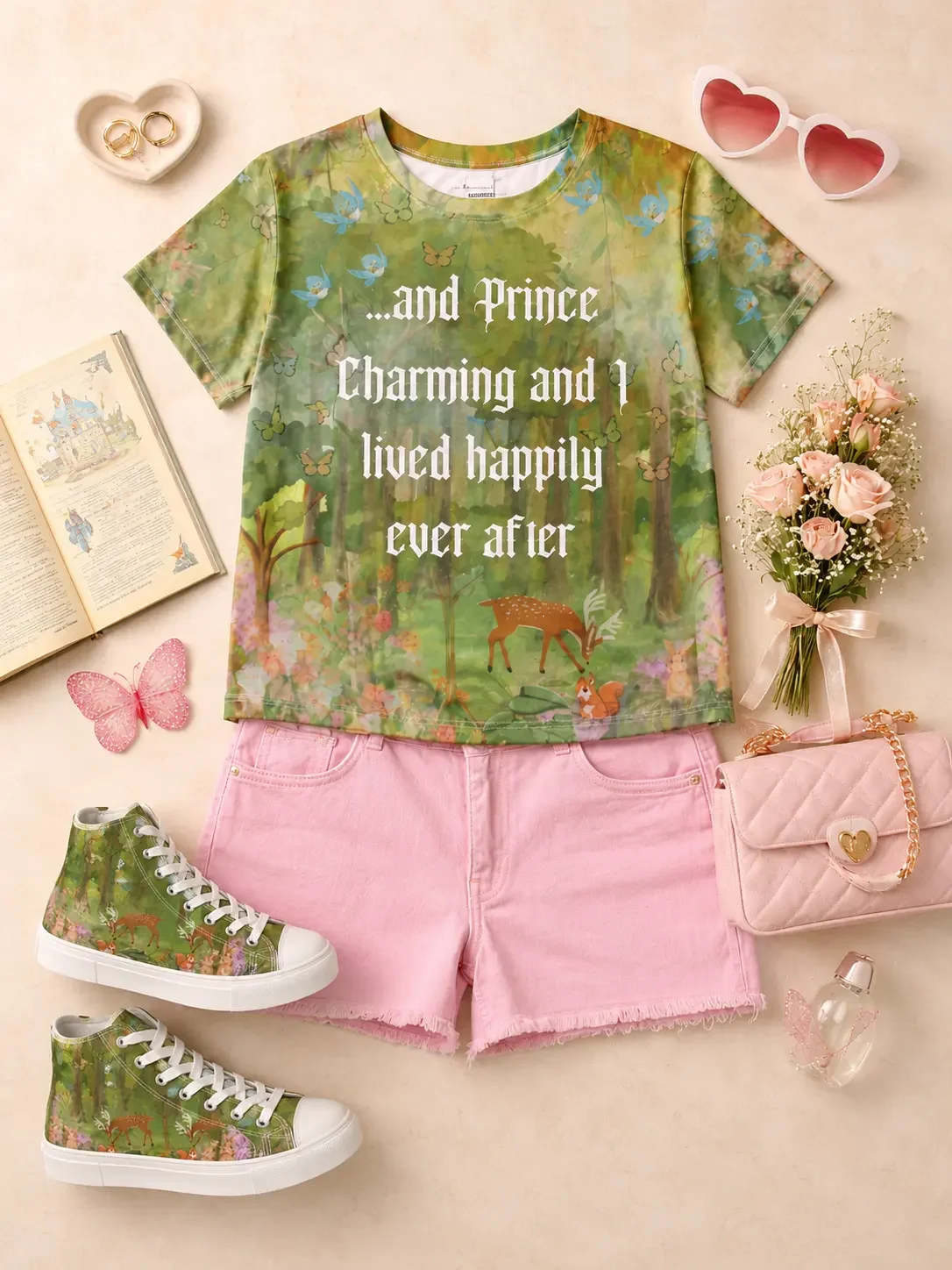 Flat lay of a green "...and Prince Charming" forest graphic tee and pink denim shorts styled with a quilted pink handbag, white sneakers, heart-shaped sunglasses, roses, and a fairytale book.