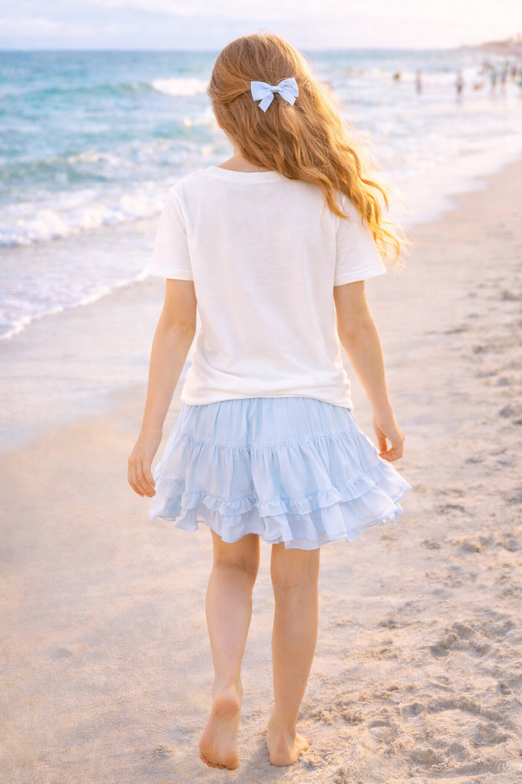 Back view of girl walking along shoreline wearing white Bahamas graphic t-shirt and blue skirt.