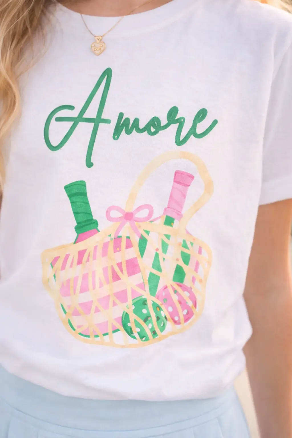 Close-up of a white graphic tee showing green "Amore" script above a pink and green pickleball paddle basket design.