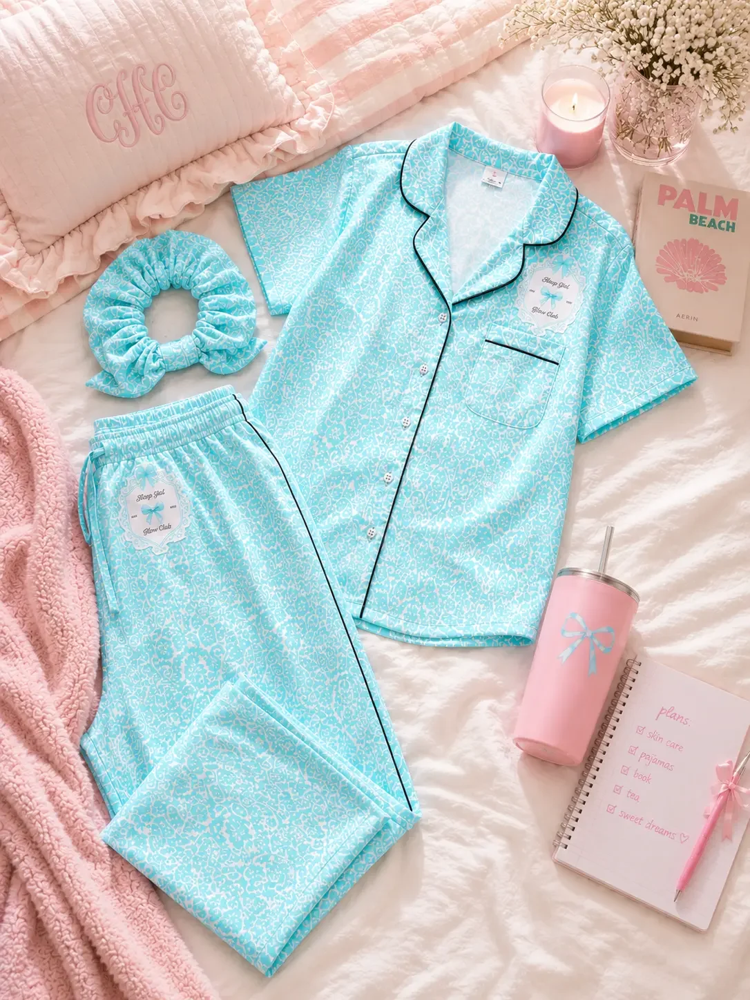 Flat lay display of a mint green patterned pajama shirt and pants with black piping, matching scrunchie, bow tumbler, and a notebook on a bed.