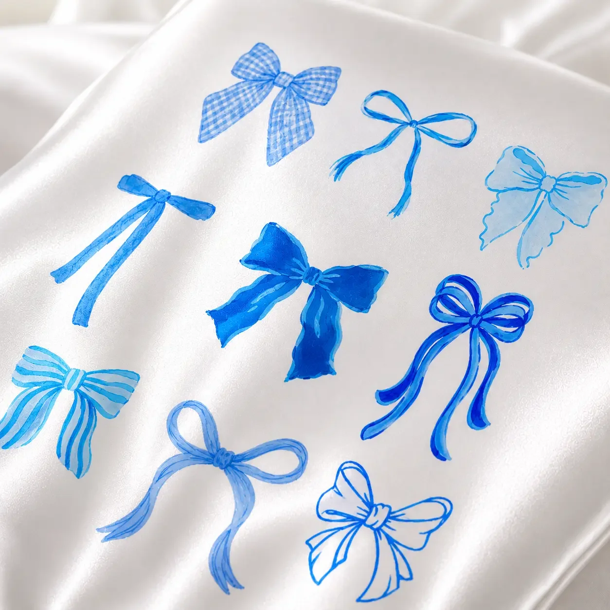 An angled macro shot of a white satin material printed with detailed shades of blue coquette ribbon bows and checkered patterns.