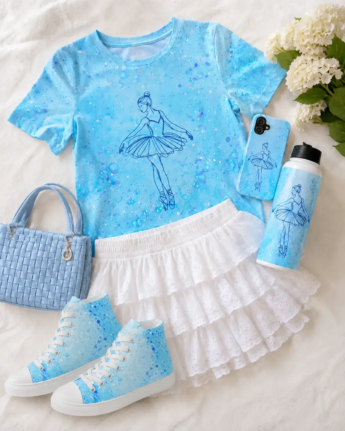 Flat lay featuring a blue ballerina graphic t-shirt styled with a tiered white ruffle skirt, light blue woven handbag, matching phone case, insulated water bottle, shoes, and white hydrangeas.