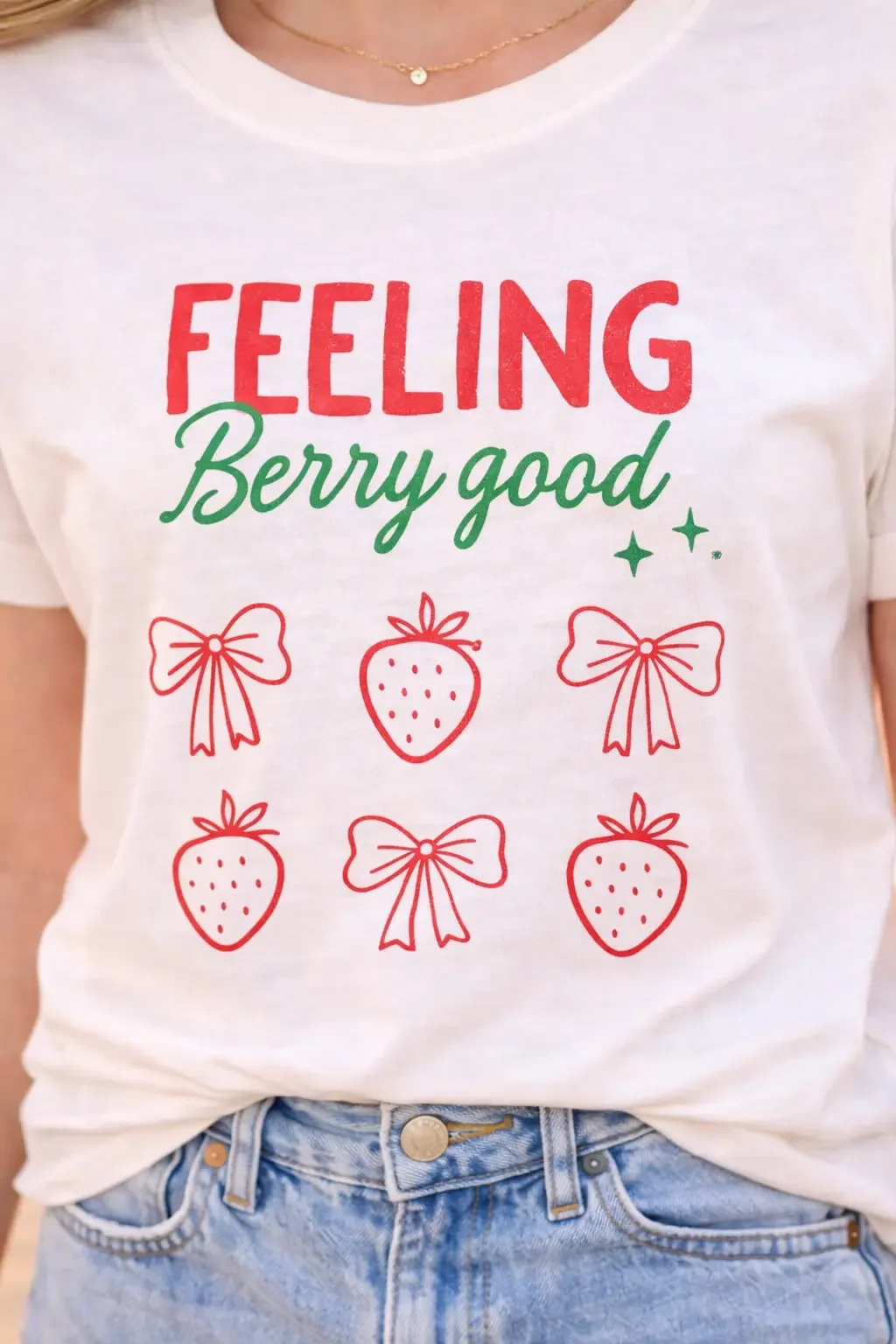 Close-up of a white graphic tee featuring the red block phrase "FEELING" above green cursive text reading "Berry good" and a grid of red strawberry and ribbon bow illustrations.