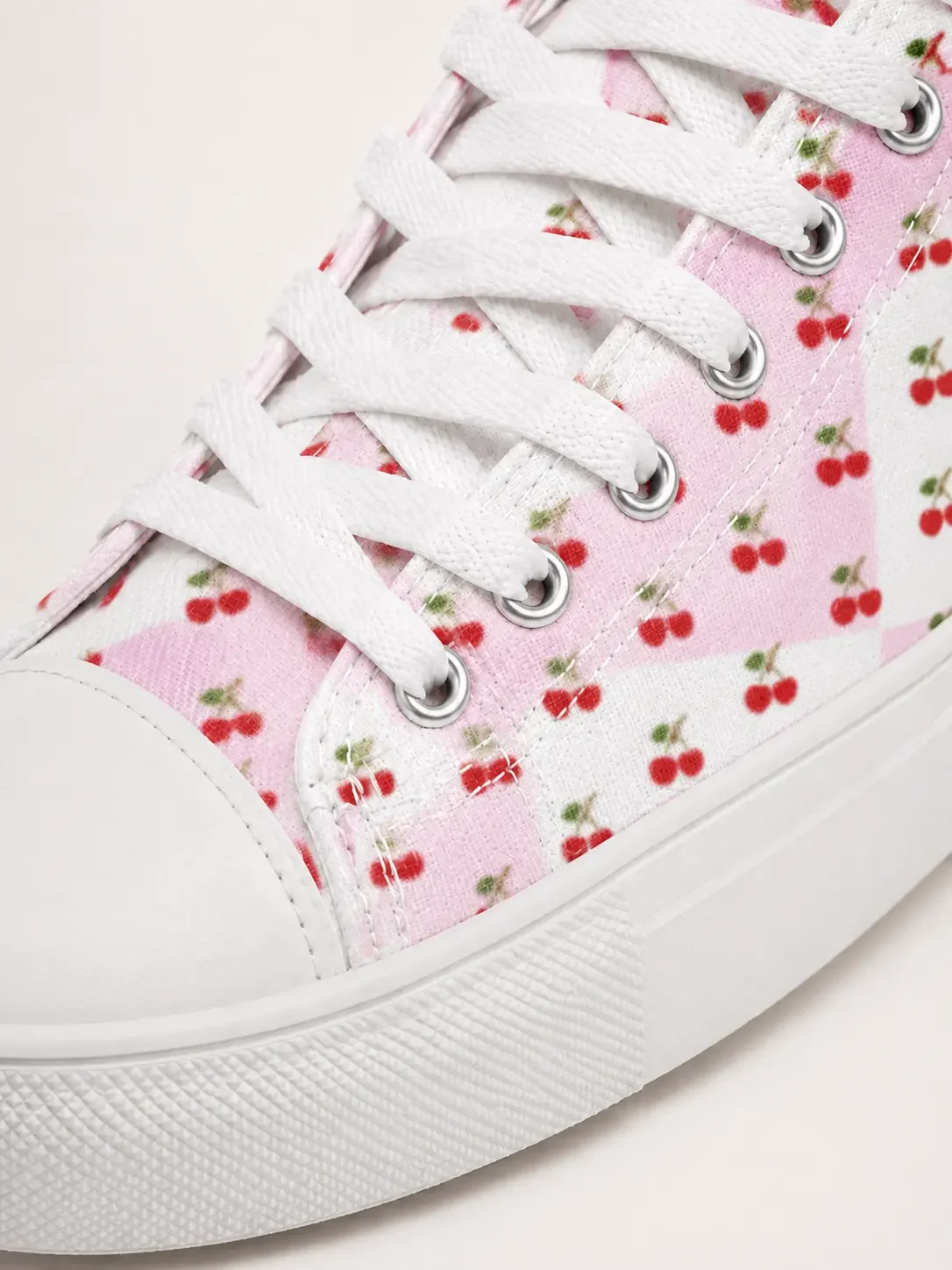 Close-up detail of white shoelaces, a white rubber sole, and a pink checkered canvas material with a cherry pattern.