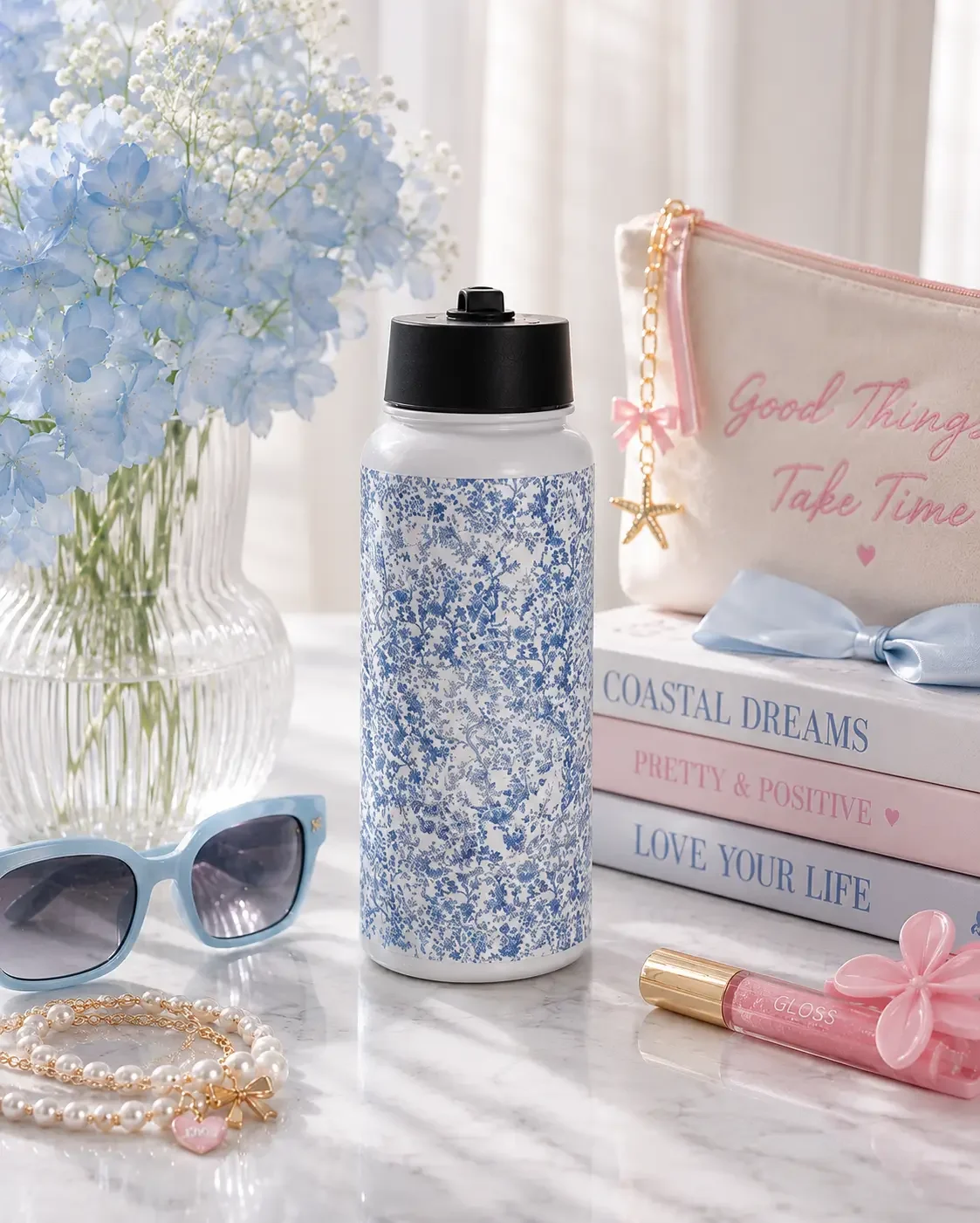A blue floral water bottle sits on marble near blue flowers, light blue sunglasses, stacked books, and a pink pouch.