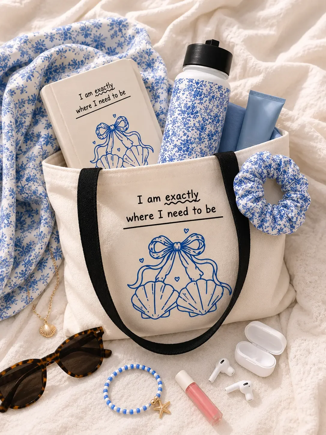 White seashell graphic tote bag packed with a blue journal, floral water bottle, scrunchie, and tech accessories.