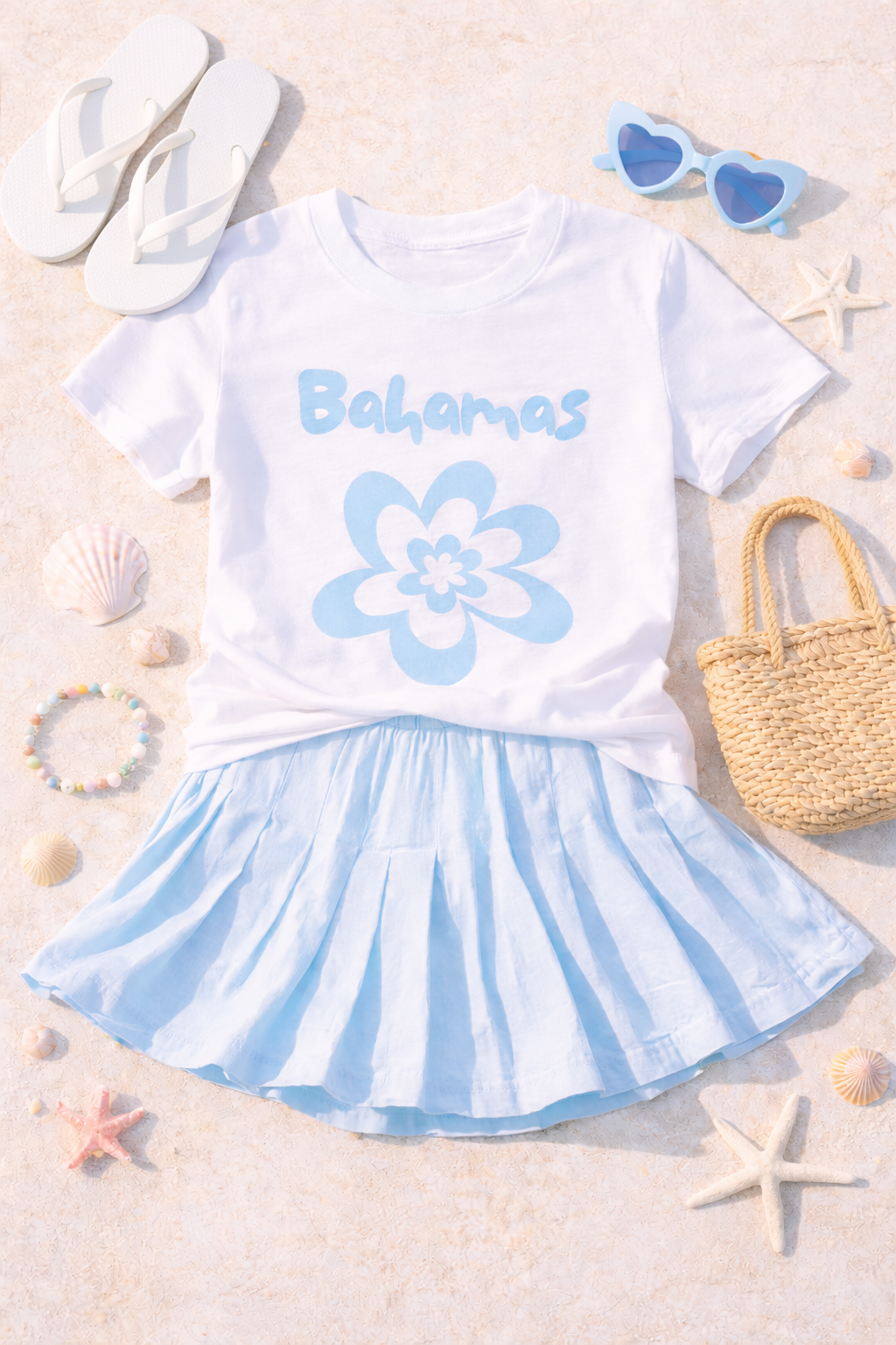 Flat lay of white Bahamas flower graphic tee styled with blue skirt, sunglasses, sandals and straw bag.