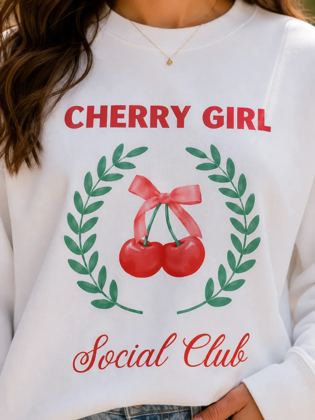 Close-up macro shot of a white crewneck sweatshirt featuring a graphic print with the red text CHERRY GIRL Social Club surrounding a green wreath.