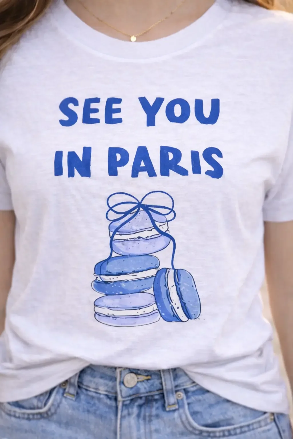 Close up of a heather grey t-shirt featuring blue see you in paris text and a stacked macaron graphic tucked into denim jeans.
