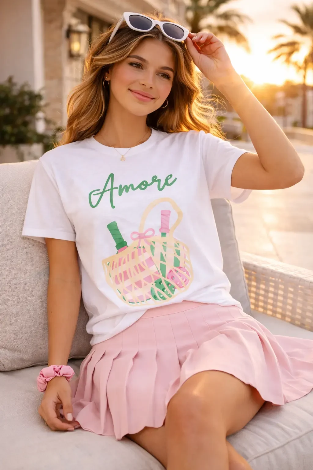Smiling young woman sitting on an outdoor lounge chair wearing a white Amore pickleball tee and a pink pleated skirt.