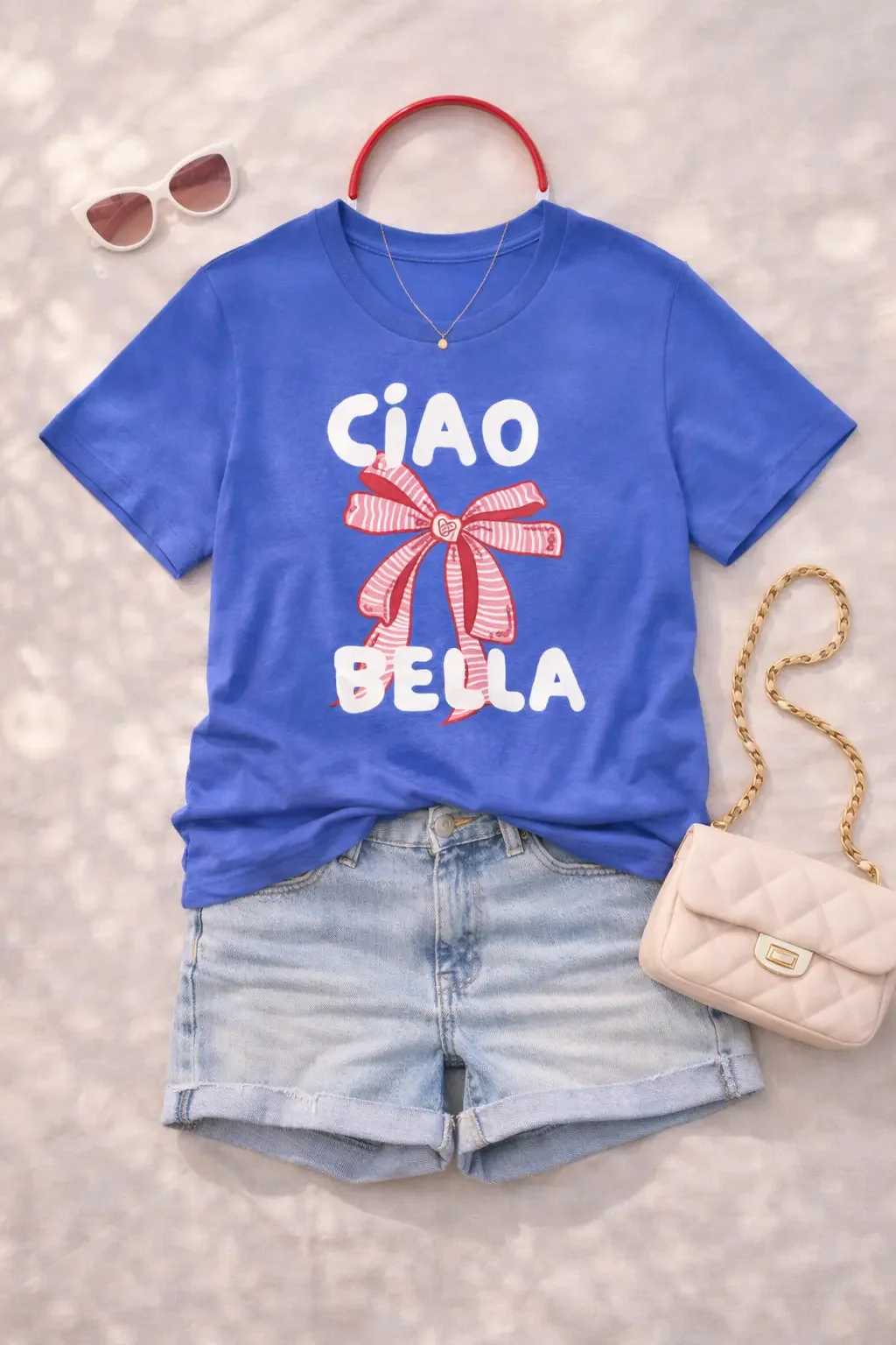 Flat lay featuring a blue Ciao Bella graphic t-shirt with a pink bow, denim shorts, and a quilted shoulder bag.