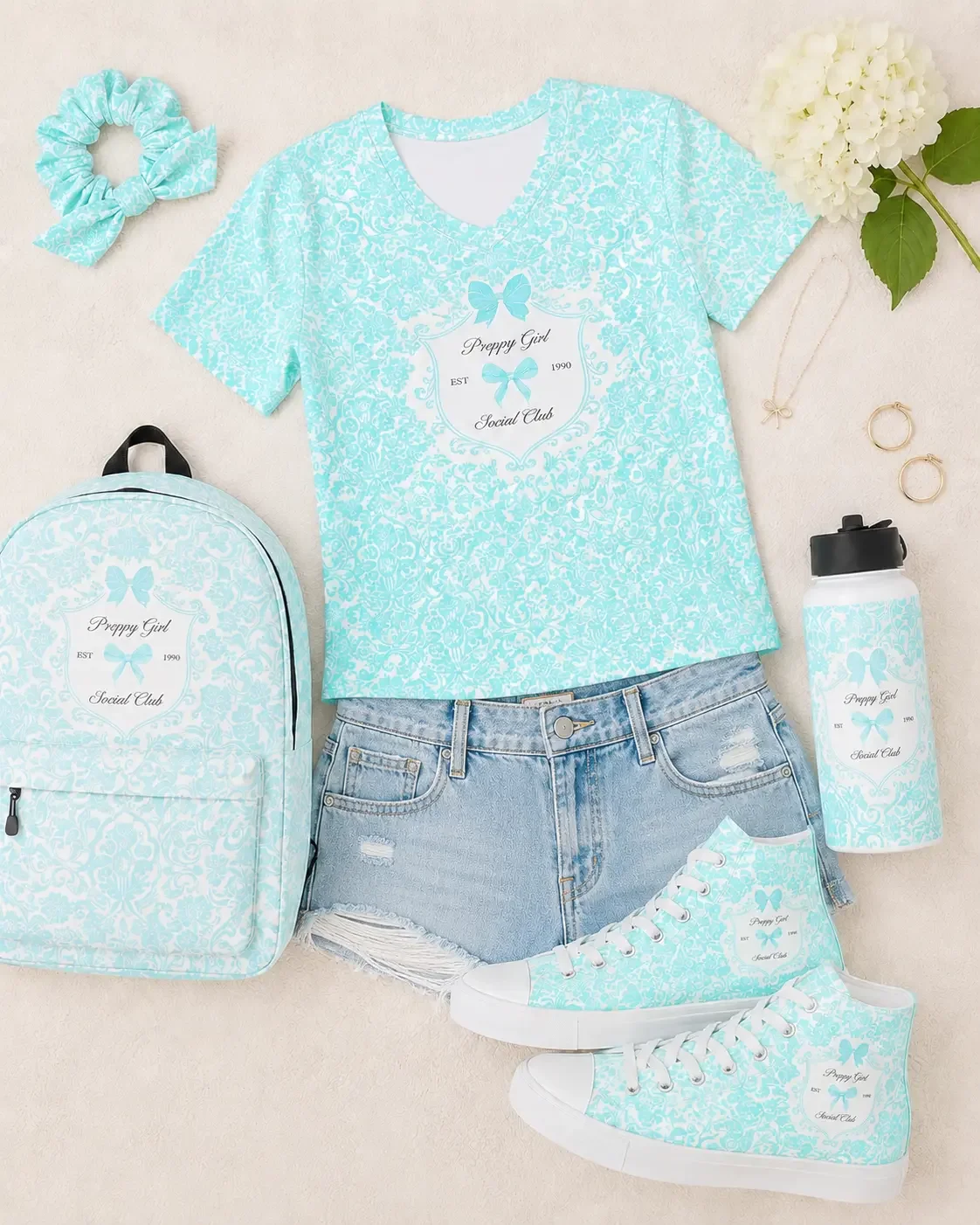 Flatlay of a blue floral bow scrunchie with a matching graphic tee, denim shorts, sneakers, water bottle, and backpack.