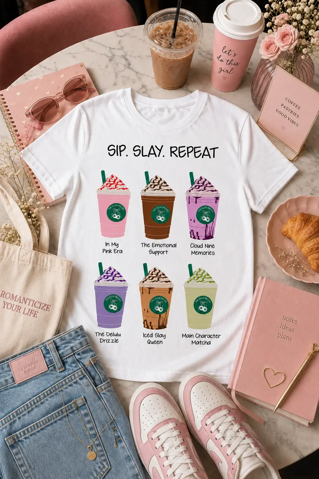 Flat lay displaying a white Sip Slay Repeat coffee graphic tee, denim shorts, pink sneakers, pink notebooks, and iced coffee.