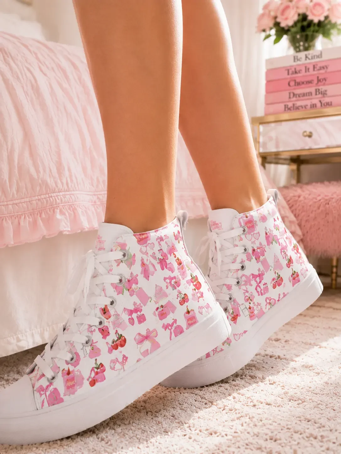 Side profile of legs wearing white high-top canvas sneakers with a pink ribbon bow and cherry pattern in a bedroom.