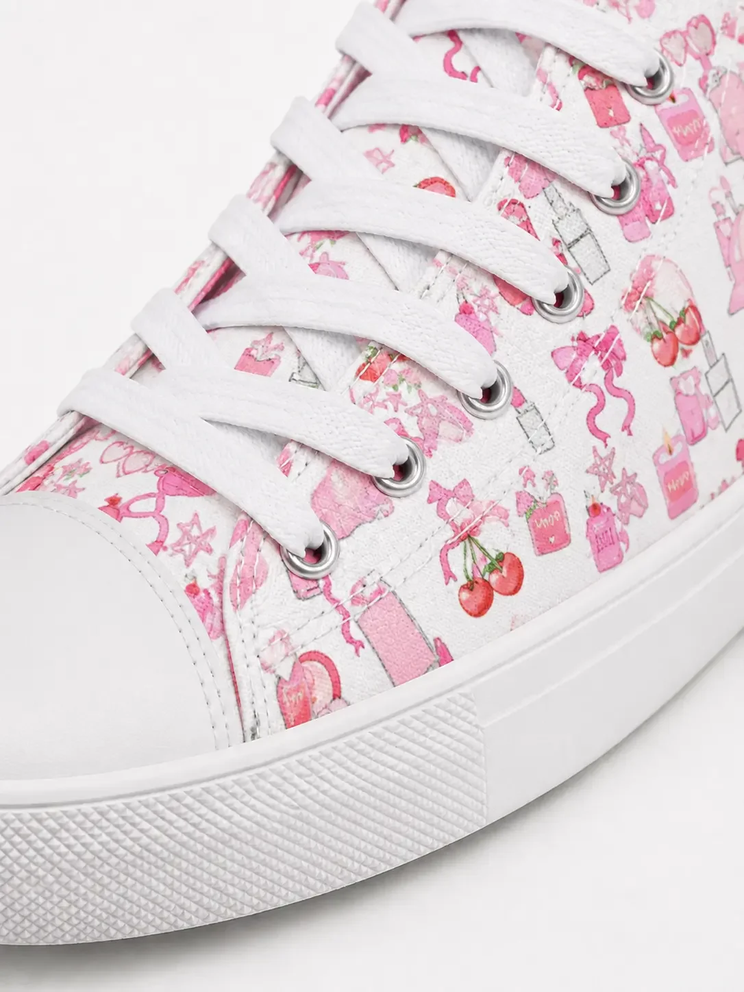 Close-up detail of white canvas shoelaces, eyelets, and a canvas material with a pink bow and cherry doodle print pattern.
