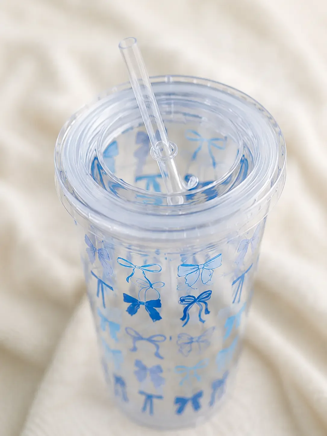 Top-down view of a clear acrylic straw tumbler featuring blue coquette ribbon bow patterns, resting on a white blanket.