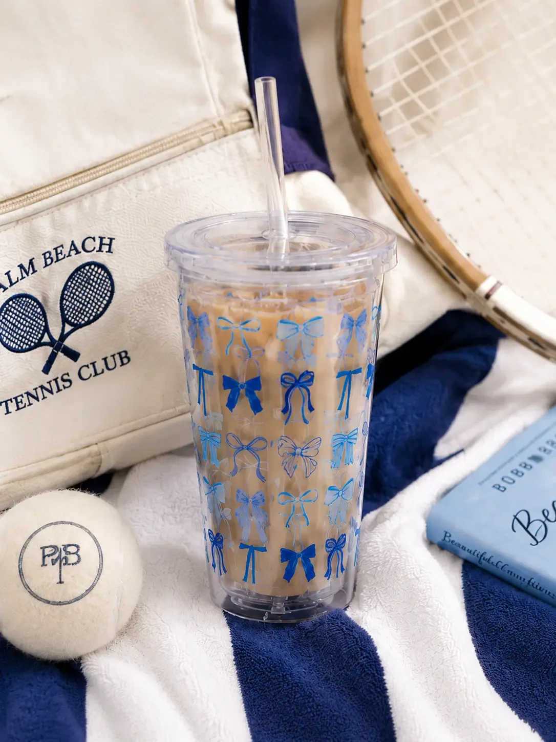 Clear insulated tumbler cup with a blue ribbon bow pattern, filled with iced coffee next to a Palm Beach tennis bag.
