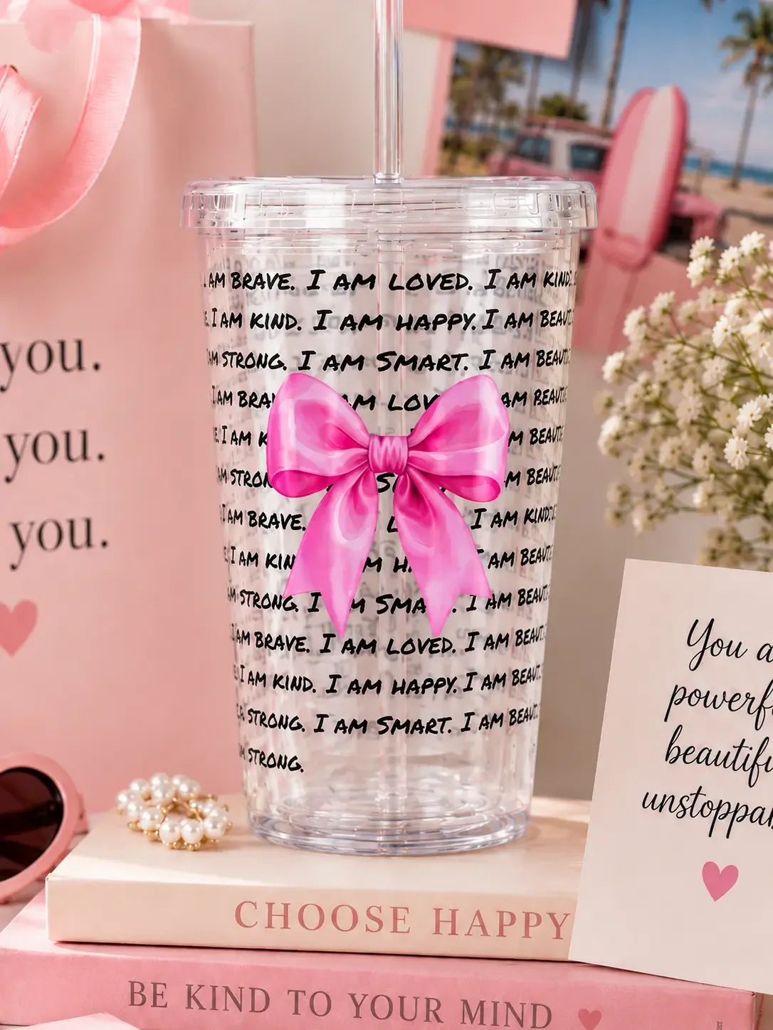 Close-up of a daily affirmation tumbler cup with a large pink bow, sitting on a "CHOOSE HAPPY" book on a desk.