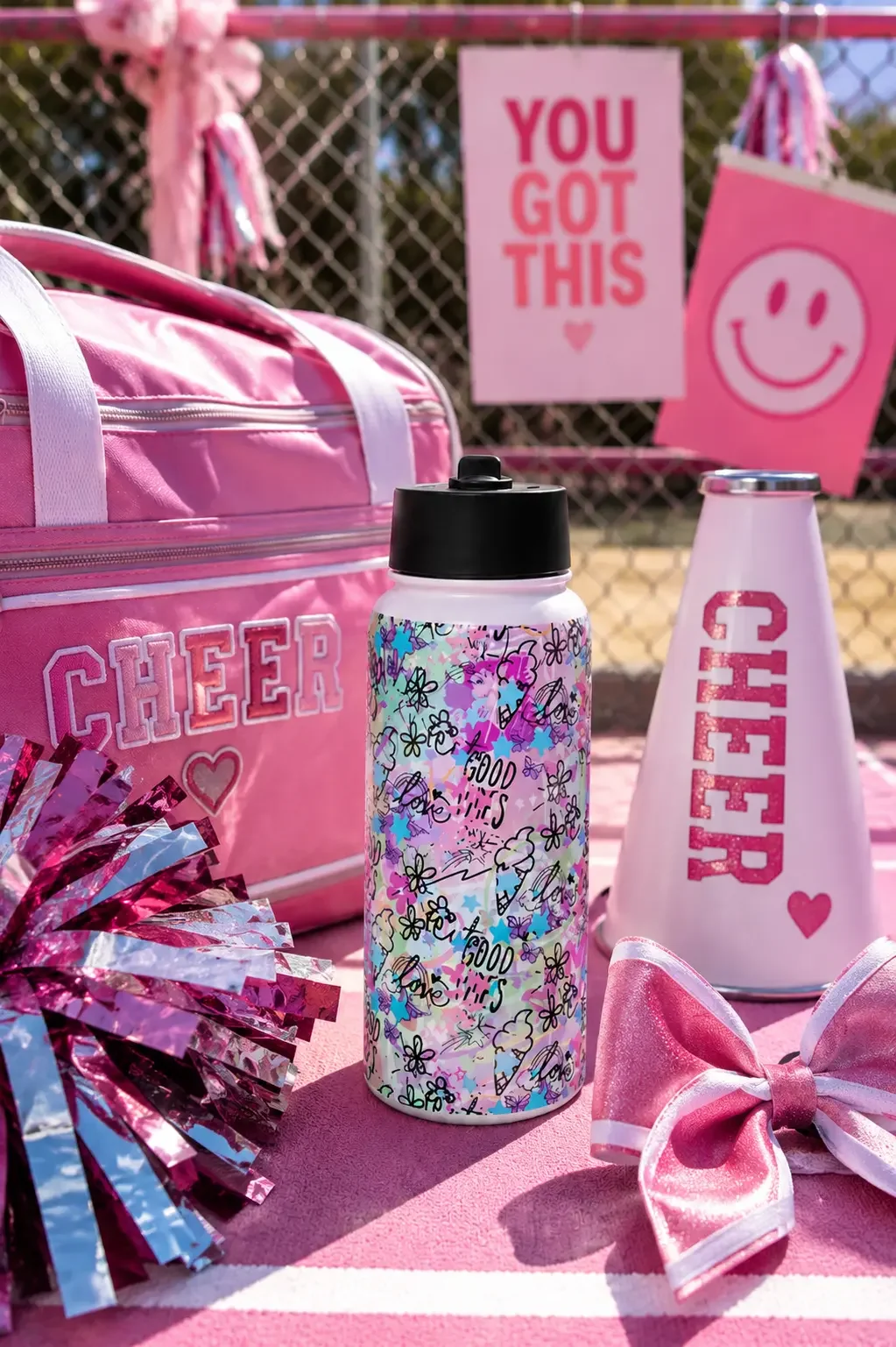 A white doodle-print water bottle sitting next to a pink cheer duffel bag, metallic pom-poms, and a megaphone.