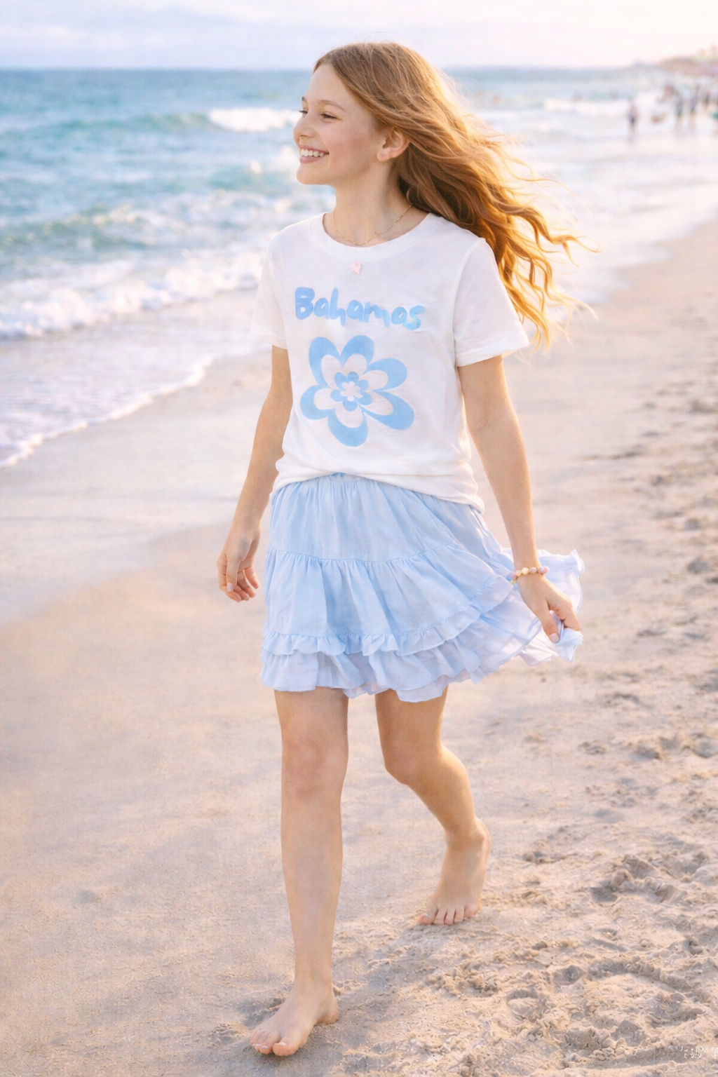 Girl smiling while walking barefoot on beach wearing white Bahamas flower graphic t-shirt.